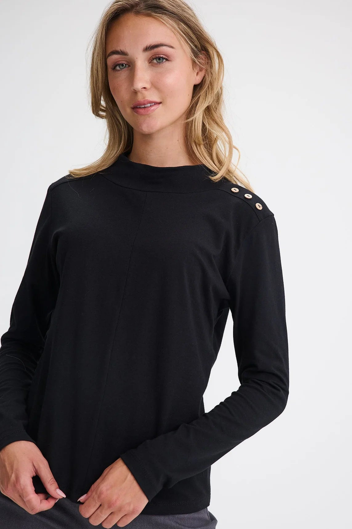 Waist up view of a woman wearing the Carleton Top by Message Factory in a Black, a long-sleeve top with button details on the shoulder. She is wearing it with grey bottoms and standing in front of a white background.