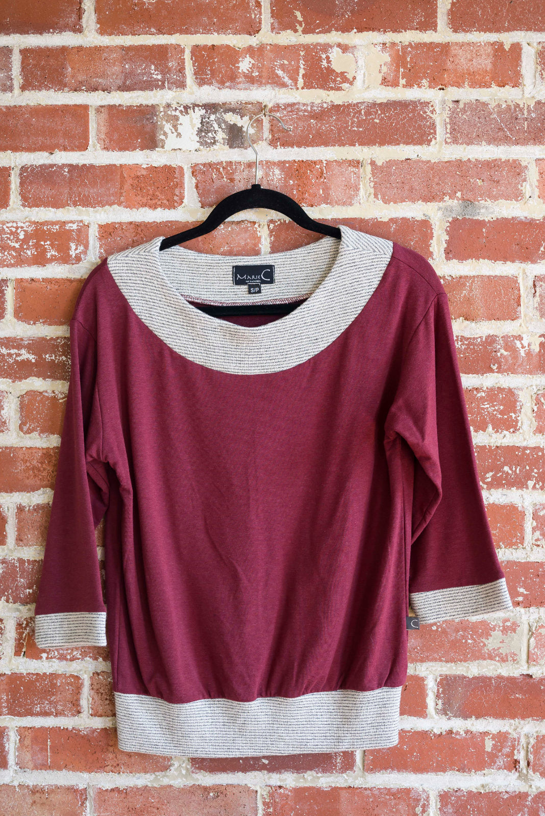 The Jaja Top by Marie C in Wine is shown against a brick wall. It's a sweatshirt with contrasting fabric bands at the wide neck, 7/8 sleeves, and hem.