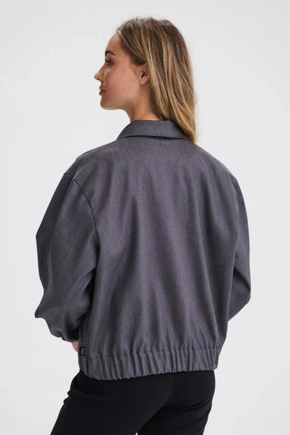 Back view of a woman wearing the Charlevoix Denim Bomber Jacket by Message Factory in Grey, with a shirt collar, button front, and large patch pockets, with black pants, standing in front of a white background