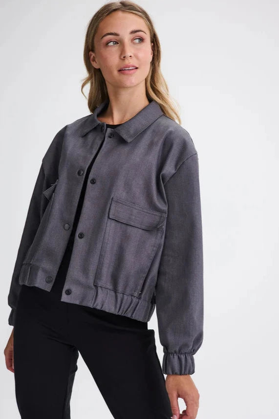 A woman wearing the Charlevoix Denim Bomber Jacket by Message Factory in Grey, with a shirt collar, button front, and large patch pockets, with black pants, standing in front of a white background