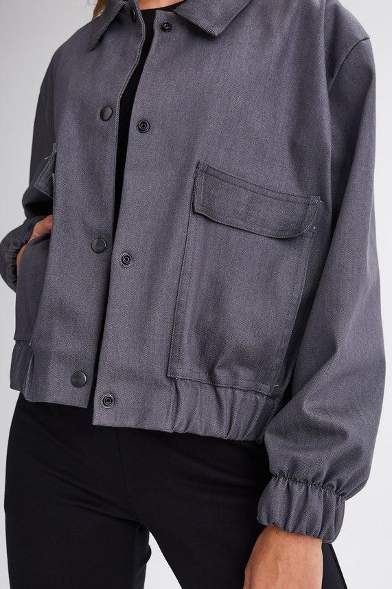 Neck down close-up of a woman wearing the Charlevoix Denim Bomber Jacket by Message Factory in Grey, with a shirt collar, button front, and large patch pockets, with black pants, standing in front of a white background