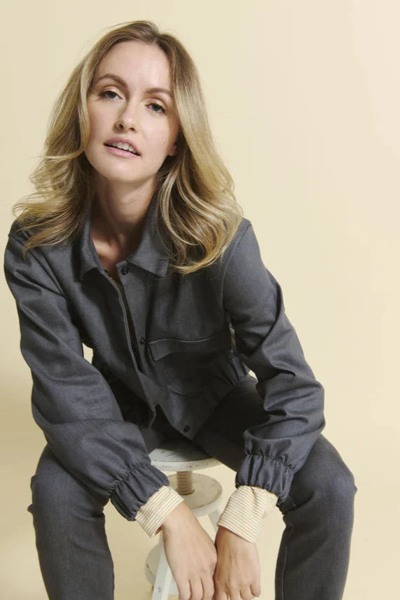 A woman wearing the Charlevoix Denim Bomber Jacket by Message Factory in Grey, with a shirt collar, button front, and large patch pockets, with grey pants, sitting in front of a beige background