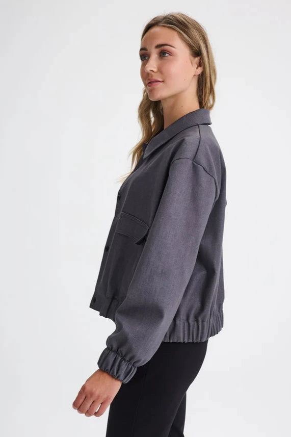 Side view of a woman wearing the Charlevoix Denim Bomber Jacket by Message Factory in Grey, with a shirt collar, button front, and large patch pockets, with black pants, standing in front of a white background
