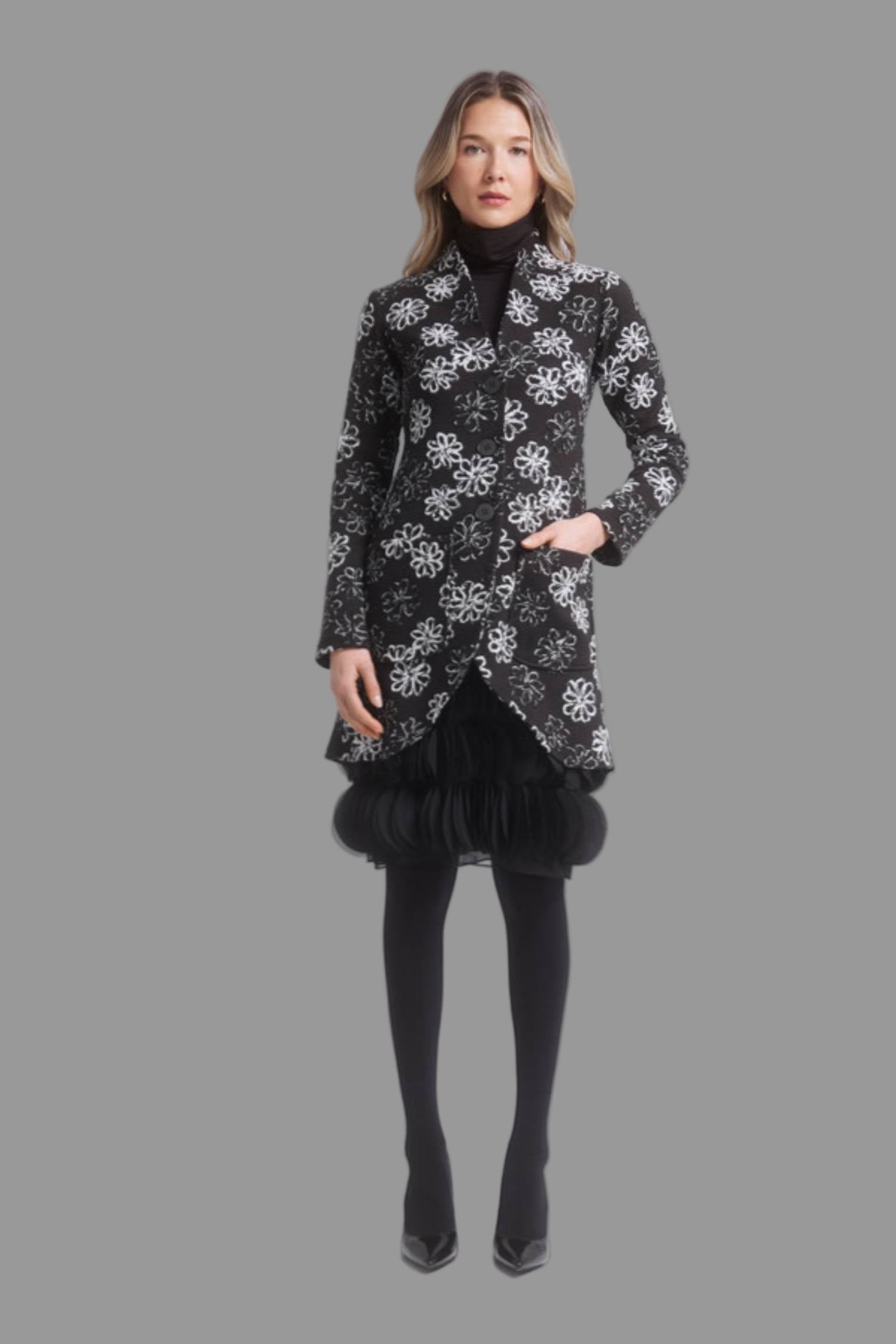A woman wearing the Laurie Jacket by Dinh Ba in Black and White Floral, a thigh-length coat with V-neck, button front, patch pocket and curved front hem. She is wearing it with a black dress and standing in front of a grey background.