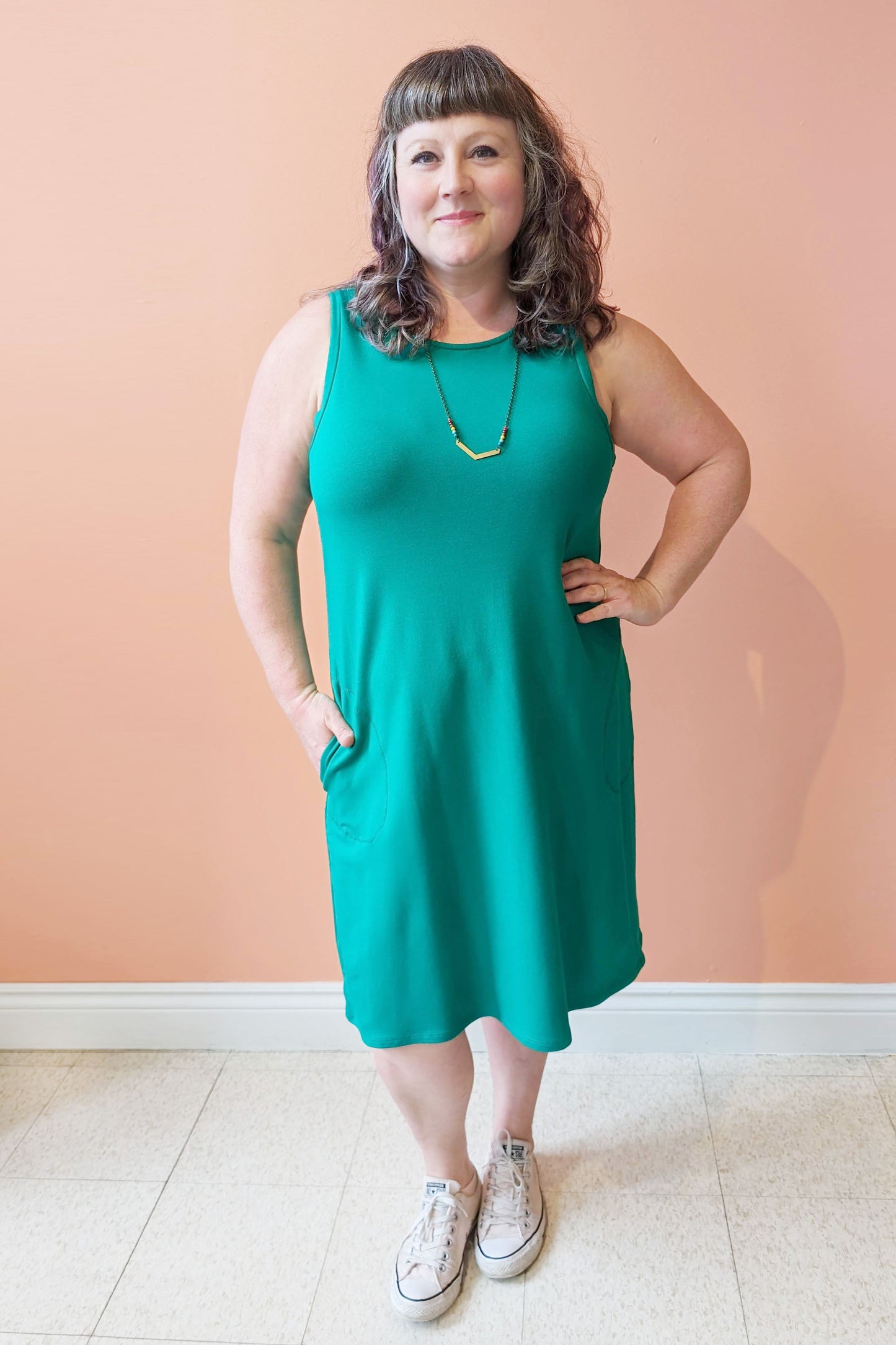 Allana is wearing the Cheri Dress by Pure Essence in Kelly Green, standing in front of a coral wall
