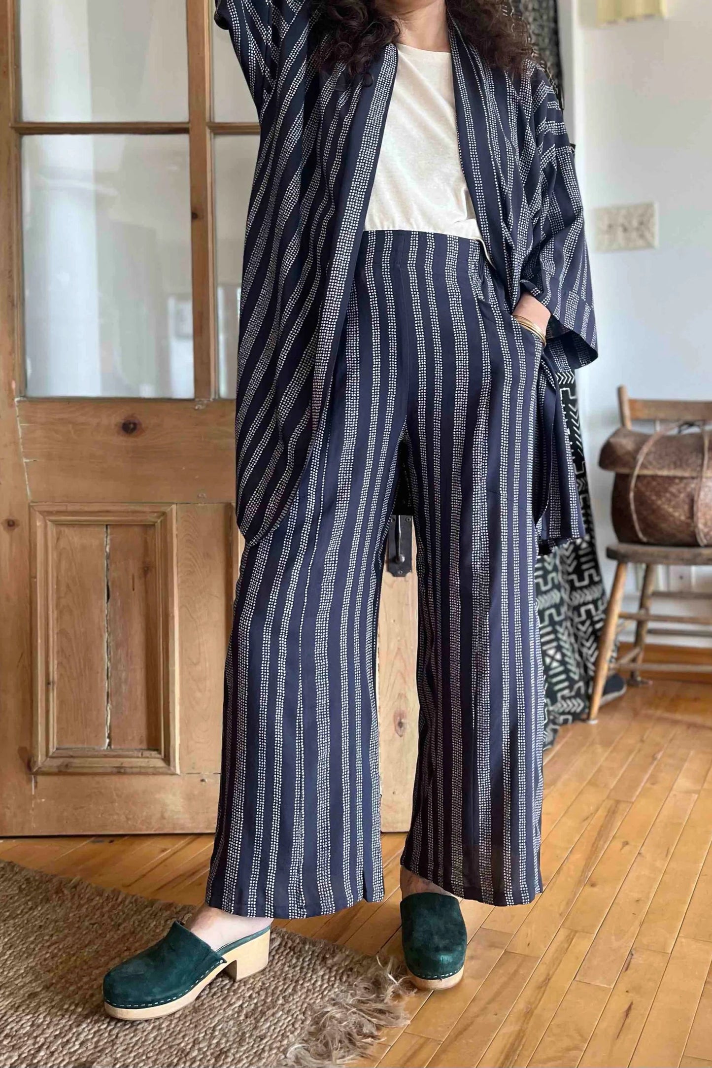 A woman wearing the Mirano Jacket by Kazak in Charcoal Dots print, an open Kimono-inspired jacket that falls to mid-thigh and has wide 3/4 sleeves. She is wearing it with white top top and matching pants and is standing in front of a wooden door.
