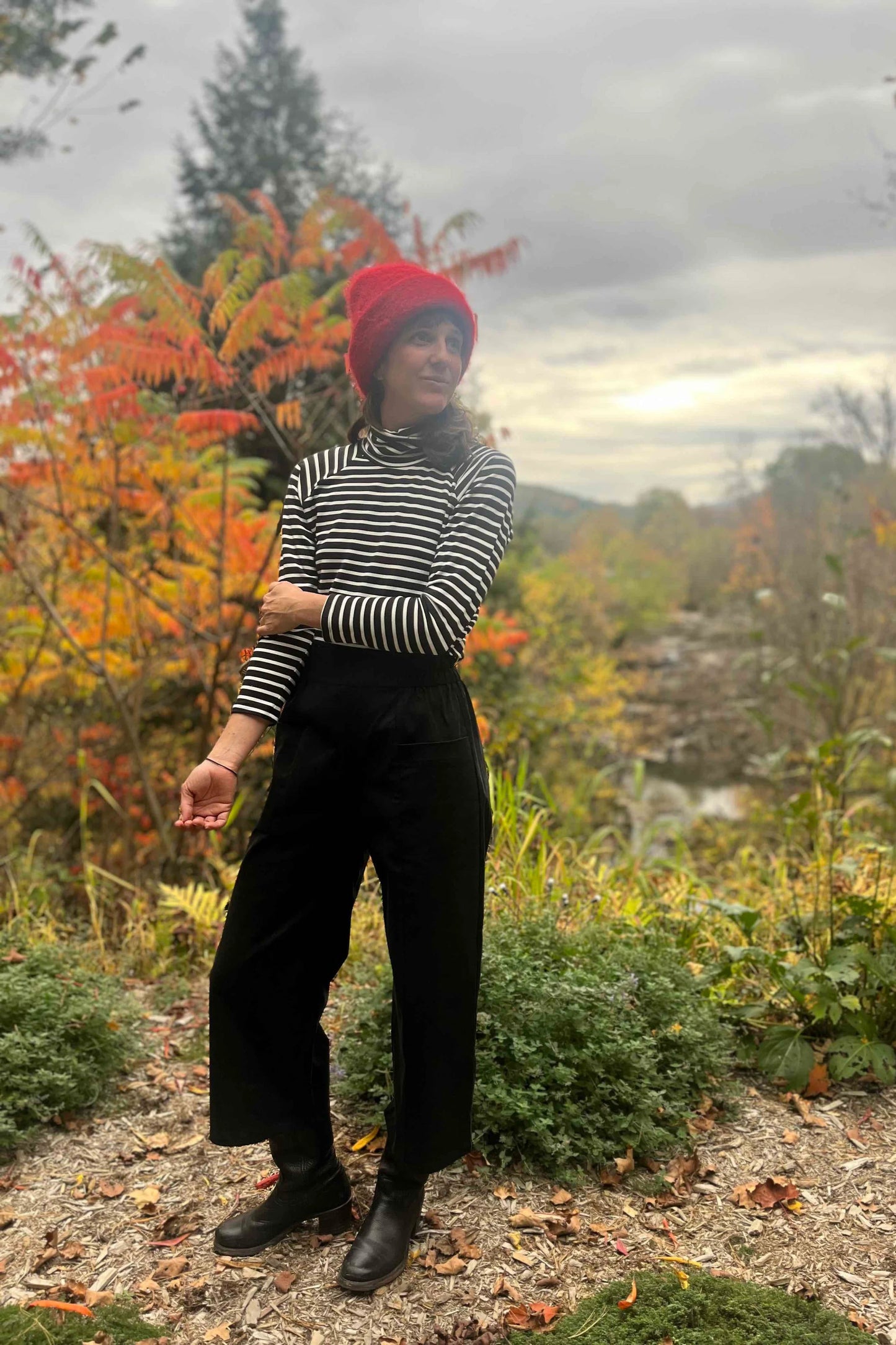 A woman wearing the Olea Pants by
Kazak in Black, wide-legged ankle-length pants with a flat-front elastic waist and large patch pockets. She is wearing them with the Oris Turtleneck in Black Stripe and standing in a forest.