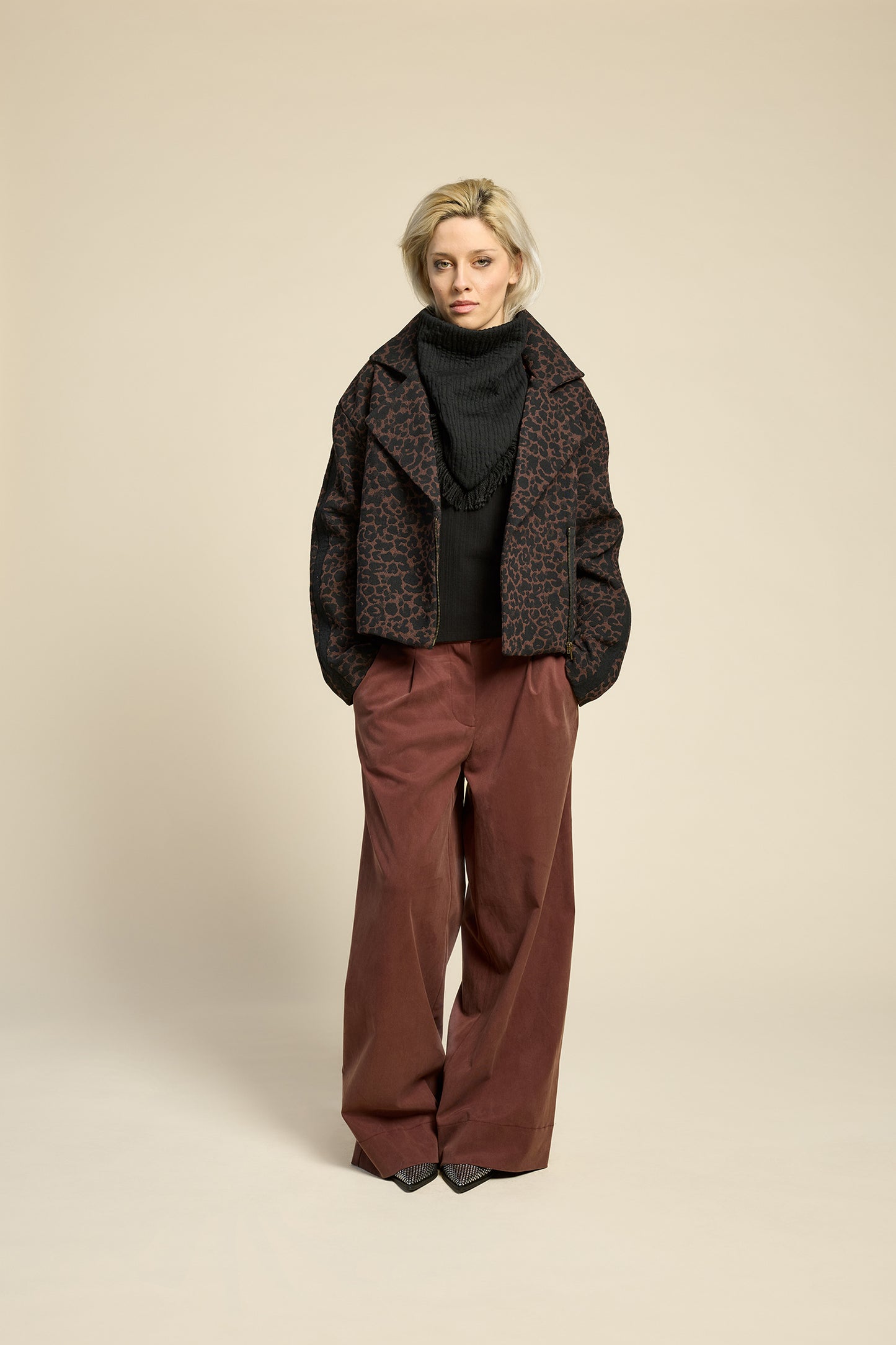A woman wearing the Perfect Day Jacket by Cokluch in Cappuccino Leopard, a motorcycle style jacket that's double breasted with a wide collar and zip front. She is wearing it with a black top and brown pants and standing in front of a beige background.