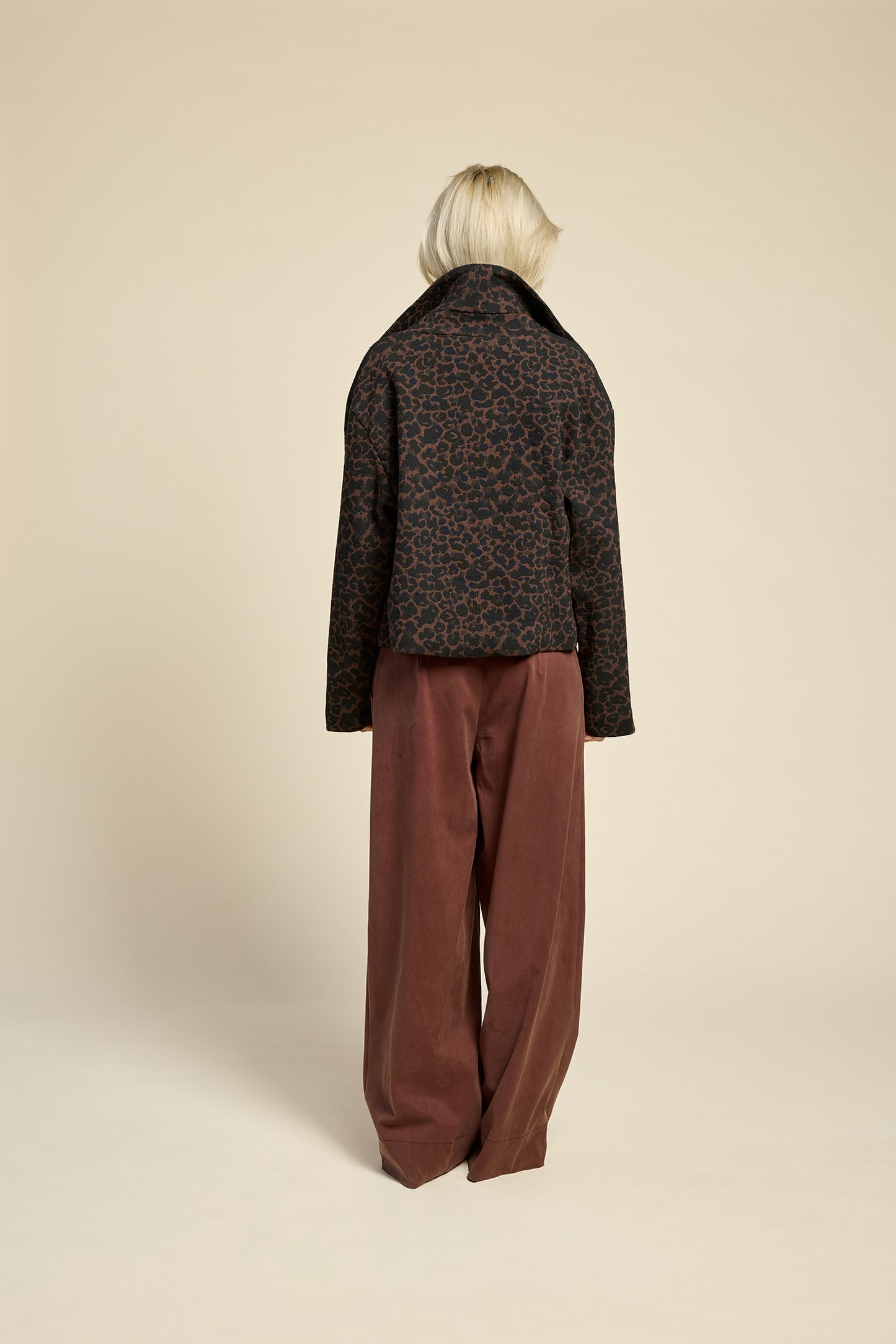 Back view of a woman wearing the Perfect Day Jacket by Cokluch in Cappuccino Leopard, a motorcycle style jacket that's double breasted with a wide collar and zip front. She is wearing it with a black top and brown pants and standing in front of a beige background.