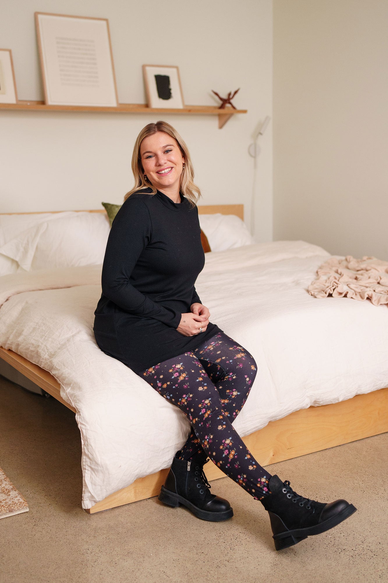 A woman wearing the Laracin Leggings by Rien ne se Perd in a small Floral print, with a black tunic. She is sitting on a bed.