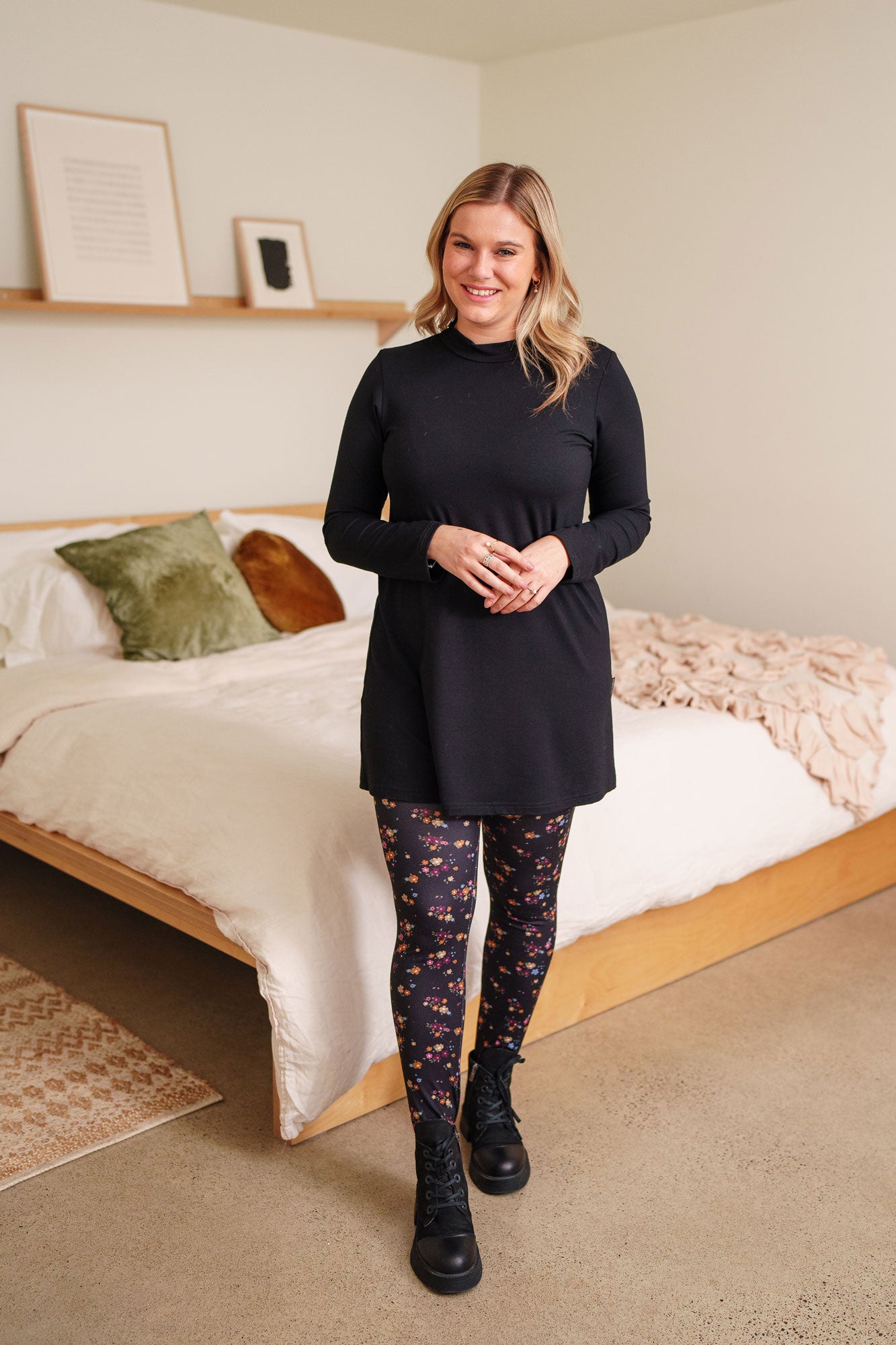 A woman wearing the Laracin Leggings by Rien ne se Perd in a small Floral print, with a black tunic. She is standing by a bed.