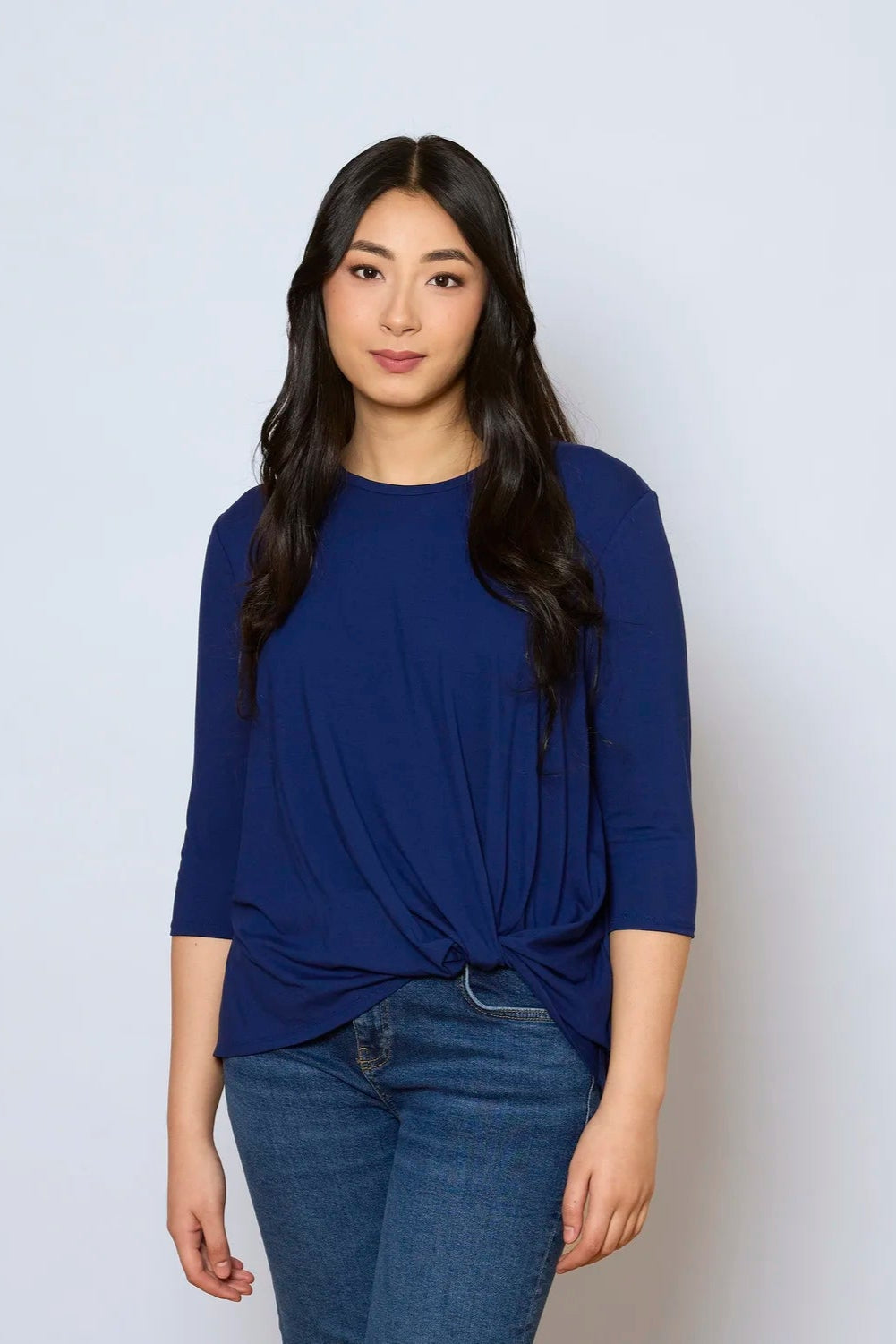 A woman wearing the Reese Top by Tangente in Blue, with a round neck, 3/4 drop sleeves, and a twisted hem in front. She is wearing it with jeans and standing in front of a white background.