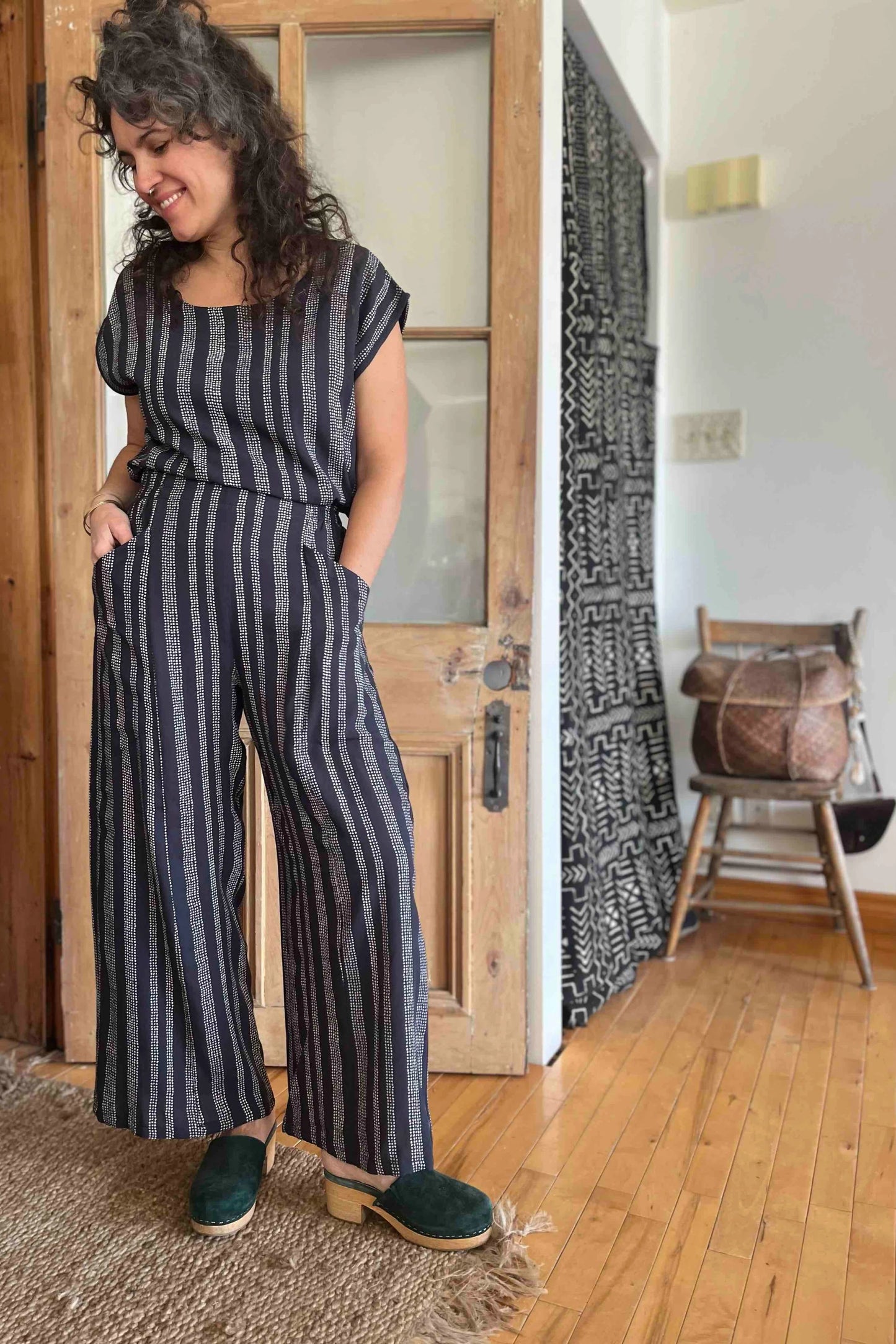 A woman wearing the Sandrigo Top by Kazak in Charcoal Dots, with short extended sleeves, a round and wide neck, and a loose fit. She is wearing it with matching pants and standing in front of a wooden door.