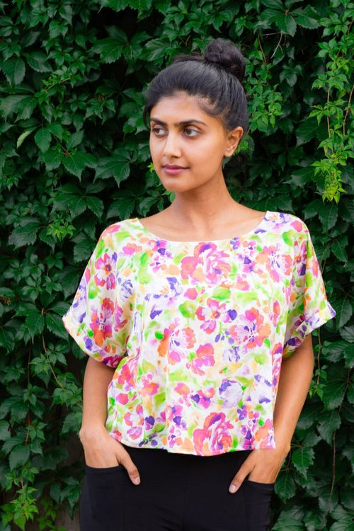 A woman wearing the Your Favourite Shirt Crop by Sara Duke in White Floral, with wide neck and rolled cuffs. She is wearing it with black pants and standing outside in front of a hedge.