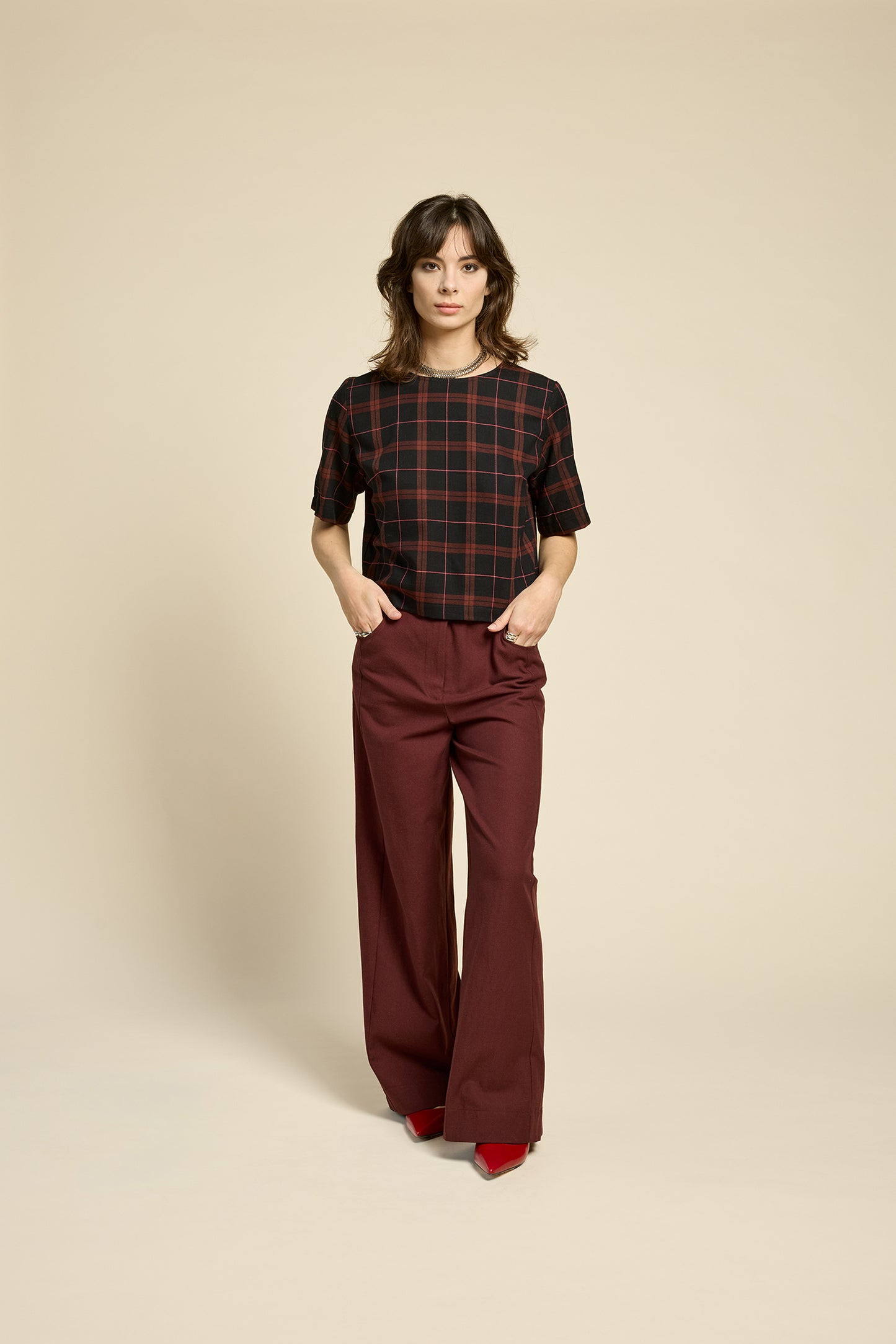 A woman wearing the Soul Love Top by Cokluch in Burgundy Plaid, with a round neck, loose fit, and elbow length sleeves. She is wearing it with with burgundy pants and standing against a beige background.
