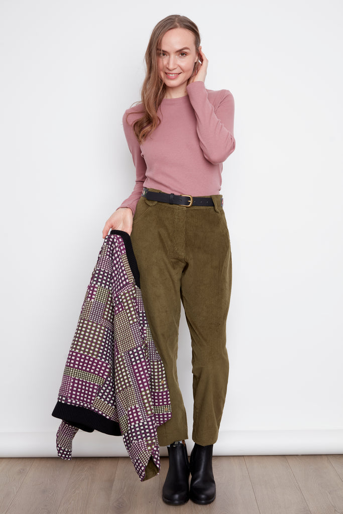 A woman wearing the Melody Mockneck Top by Julie in Rosewood, a ribbed knit top with a short cross-over collar and long sleeves. She is wearing it with the green corduroy pants and holding a patterned jacket against a white background.