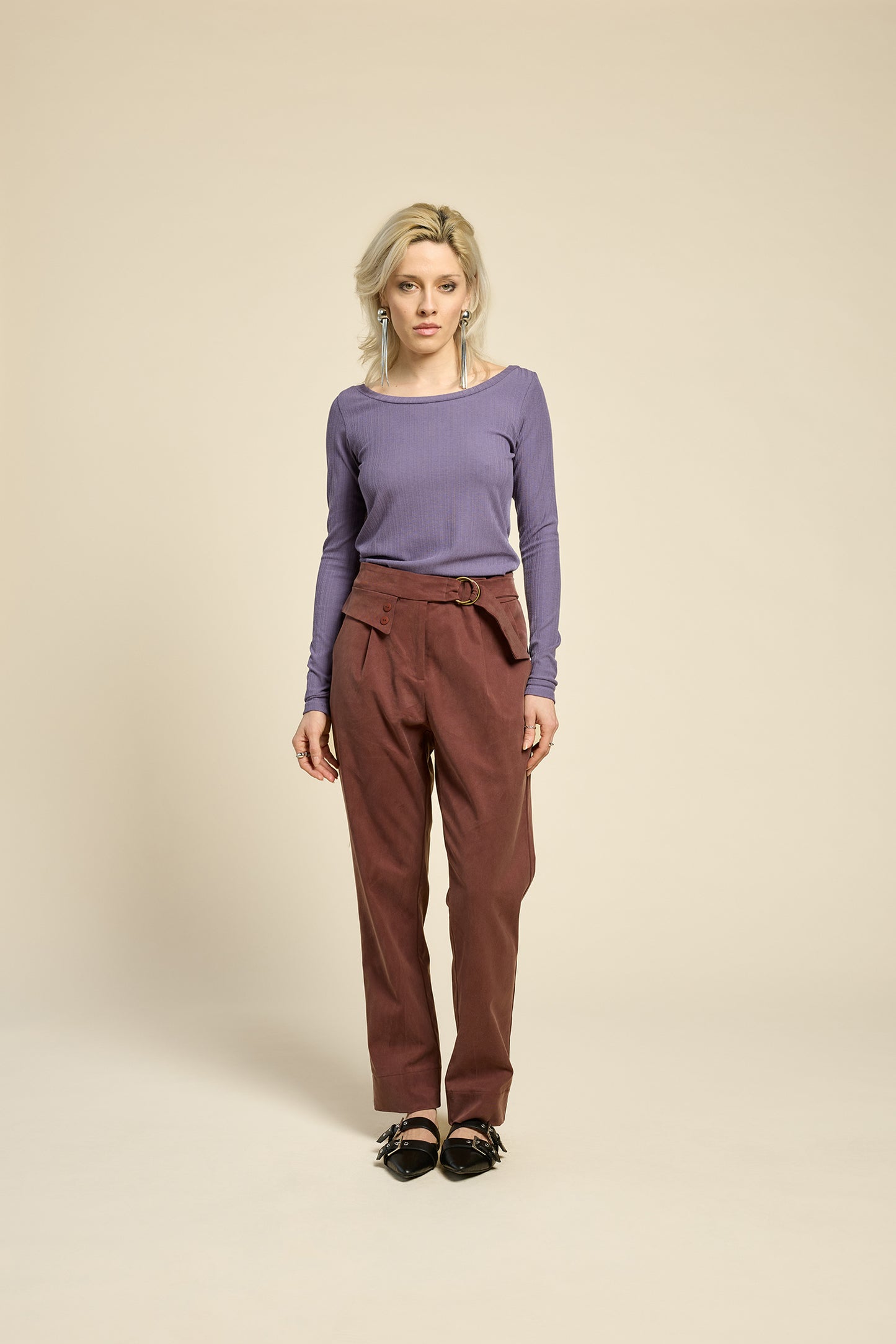 A woman wearing the Fool Pants by Cokluch in Cappucino, featuring an attached belt with D-rings, a flap and button detail at the waist, and tapered ankle-length legs. She is wearing them with a purple top and standing in front of a beige background.