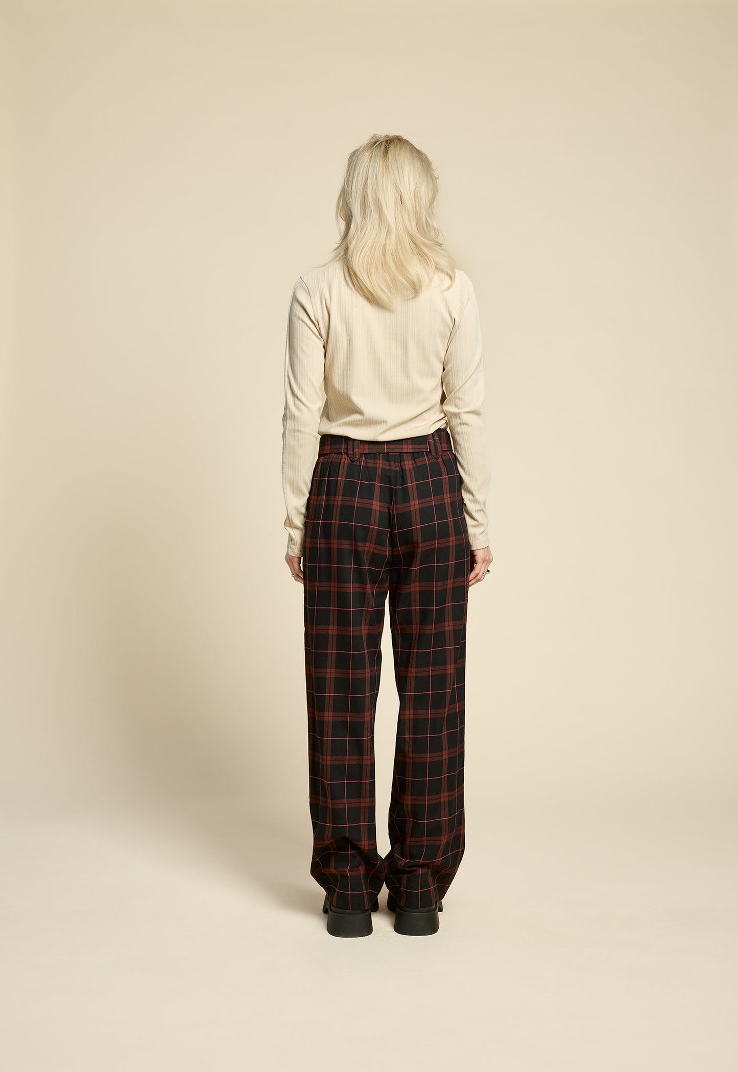 Back view of Back view of a woman wearing the West Coast Pants by Cokluch in Burgundy Plaid, featuring flared legs, an attached belt with a D-ring closure, and pointed belt loops. She is wearing them with a beige top and standing in front of a beige background.