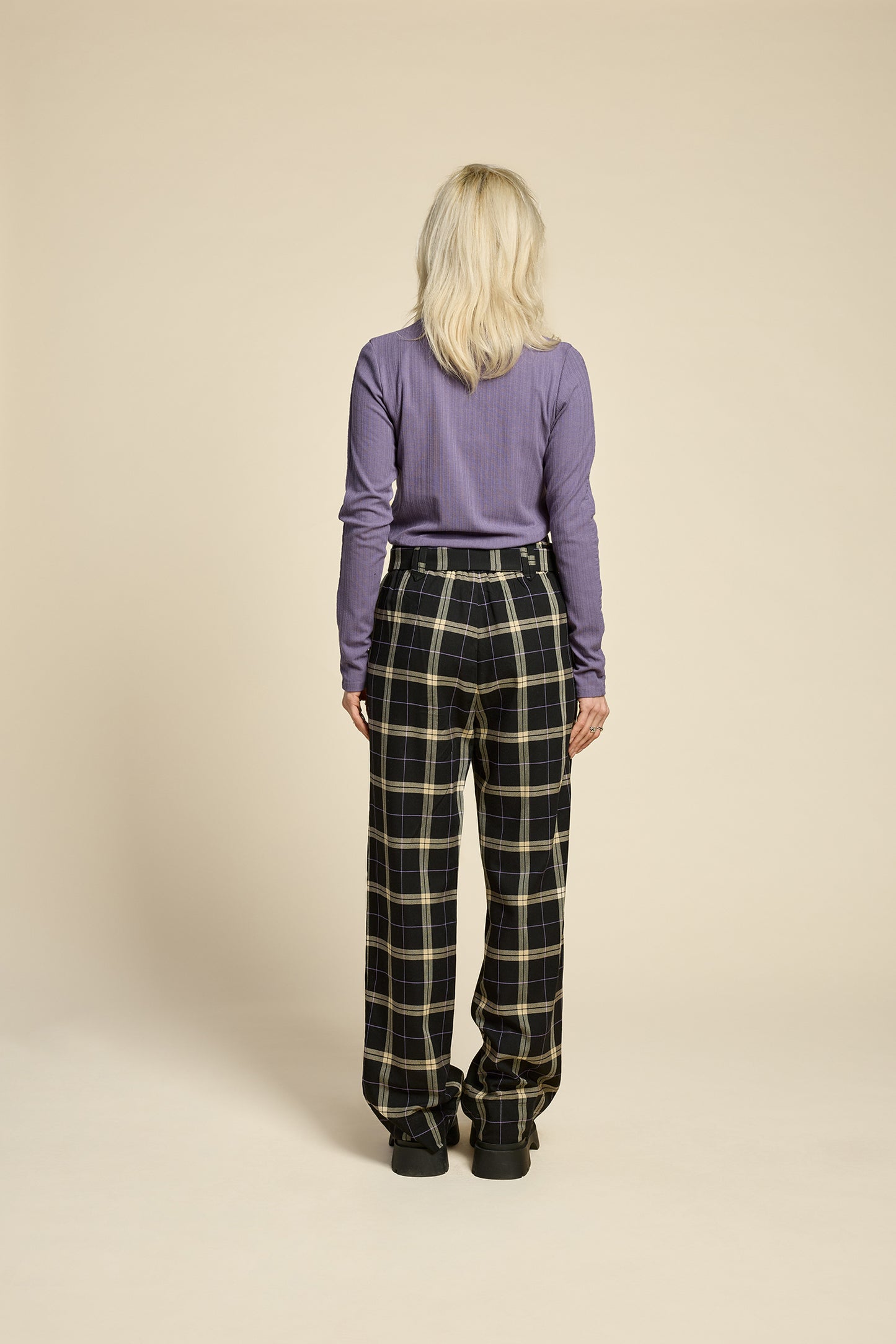 Back view of a woman wearing the West Coast Pants by Cokluch in Black Plaid, featuring flared legs, an attached belt with a D-ring closure, and pointed belt loops. She is wearing them with a purple top and standing in front of a beige background.
