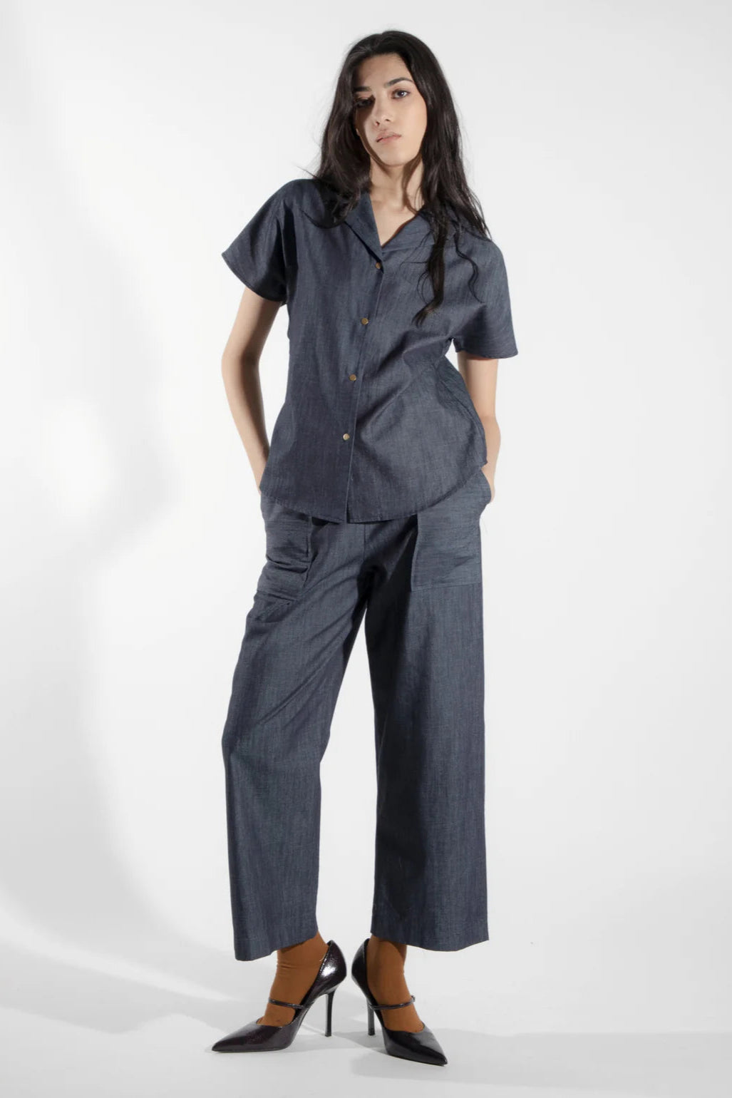 A woman wearing the Alisme Pants by Eve Lavoie in Denim, slightly cropped wide-legged pants with side patch pockets and an elastic waist. She is wearing them with a matching top and standing in front of a white background.