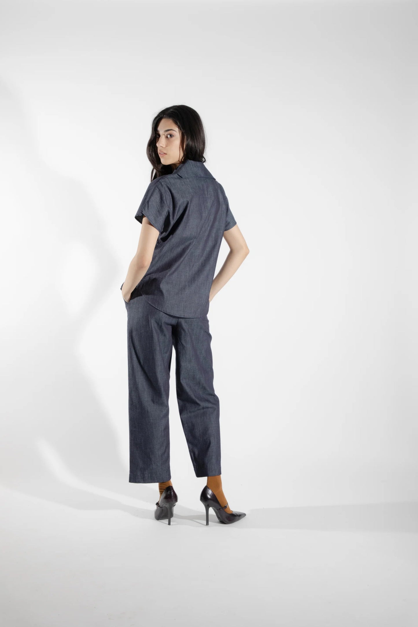 Back view of a woman wearing the Alisme Pants by Eve Lavoie in Denim, slightly cropped wide-legged pants with side patch pockets and an elastic waist. She is wearing them with a matching top and standing in front of a white background.