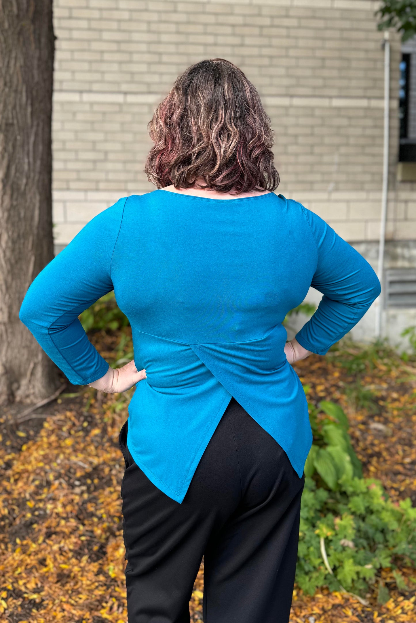 Back view of a woman wearing the Studio D Fela top in Teal, featuring 3/4 sleeves and a split hem. She is wearing black pants and standing in front of a garden.