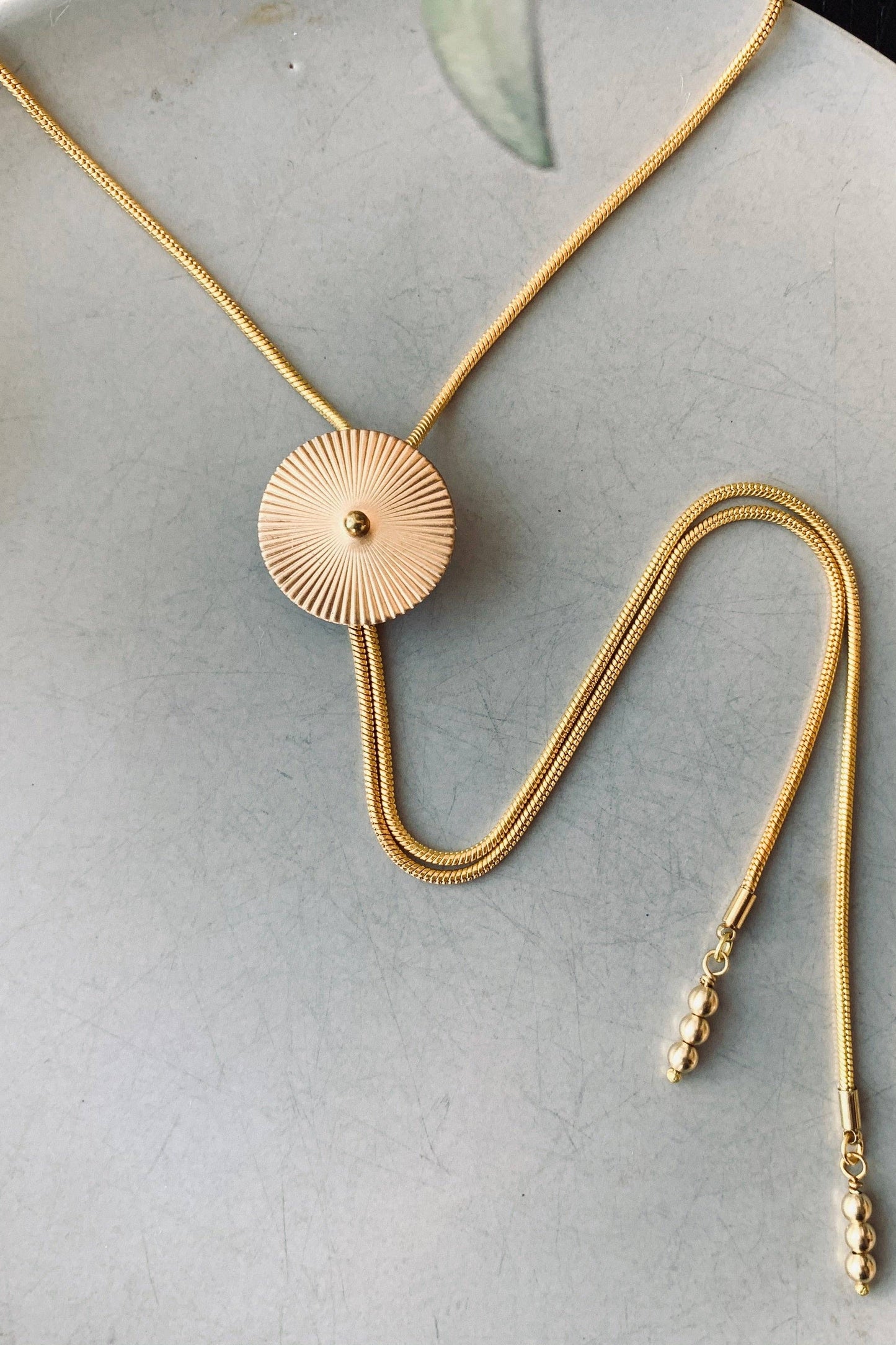 The TROLLOP Mini Bolo Tie from Lord Violet, featuring a sunburst design on a gold bolo chain, is shown against a grey background.