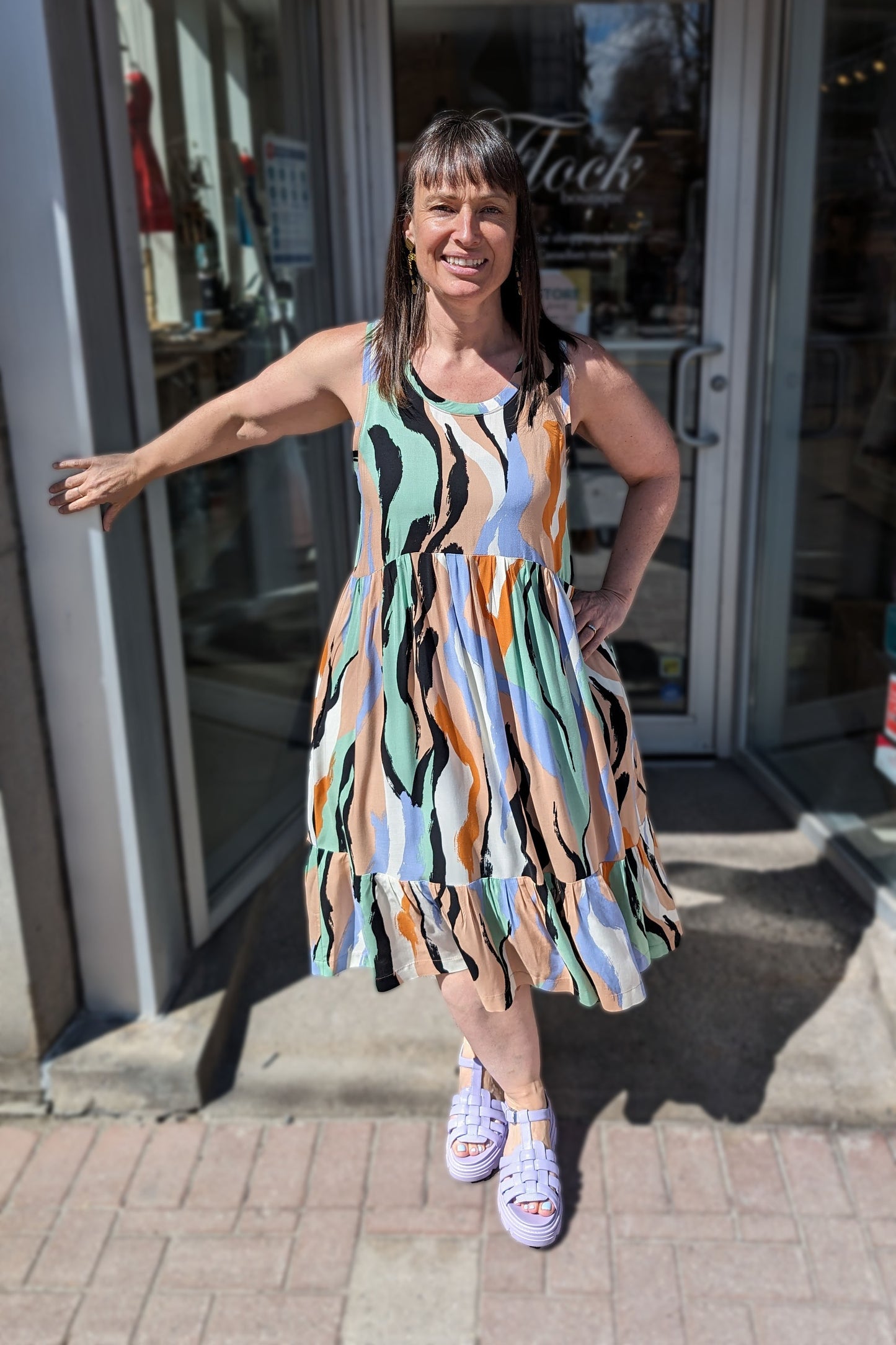 Bridget wearing the Rose Dress by Copious in Mint/Peach abstract print, a sleeveless dress with an empire waist, tiered mid-calf length skirt, and pockets. She is standing in the doorway at Flock.