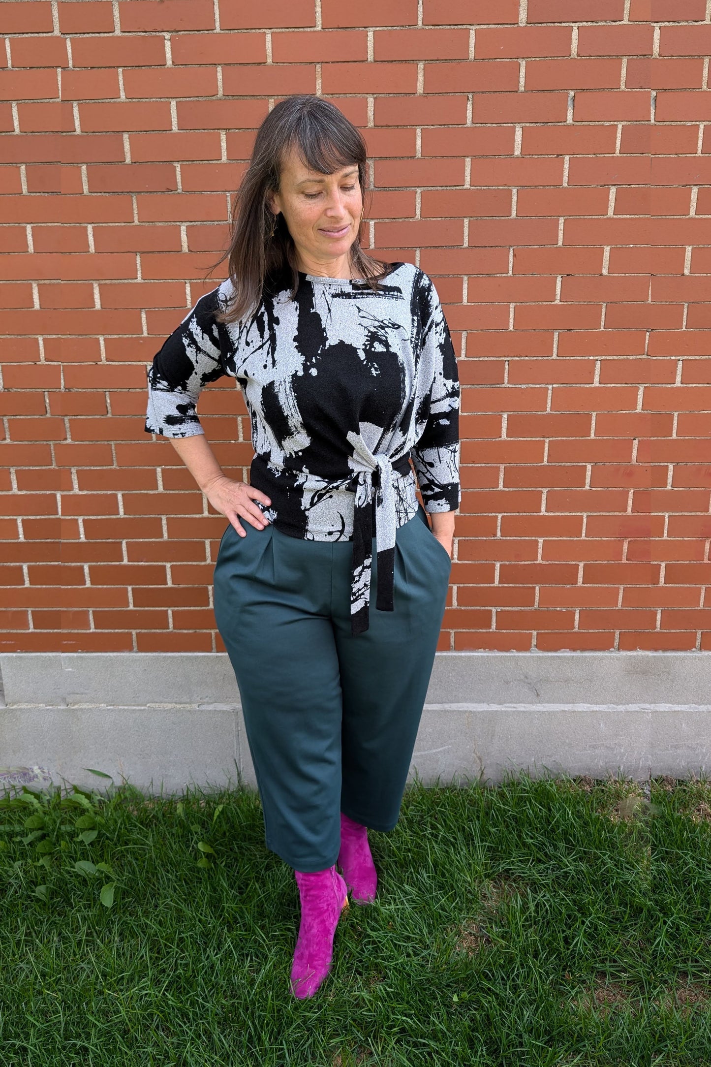 A woman wearing the NIna Top by Compli K with the Wilfred Pants by Melow in Forest - featuring
a wide pull-on waistband, front pleats, and cropped legs, standing in front of a brick wall.