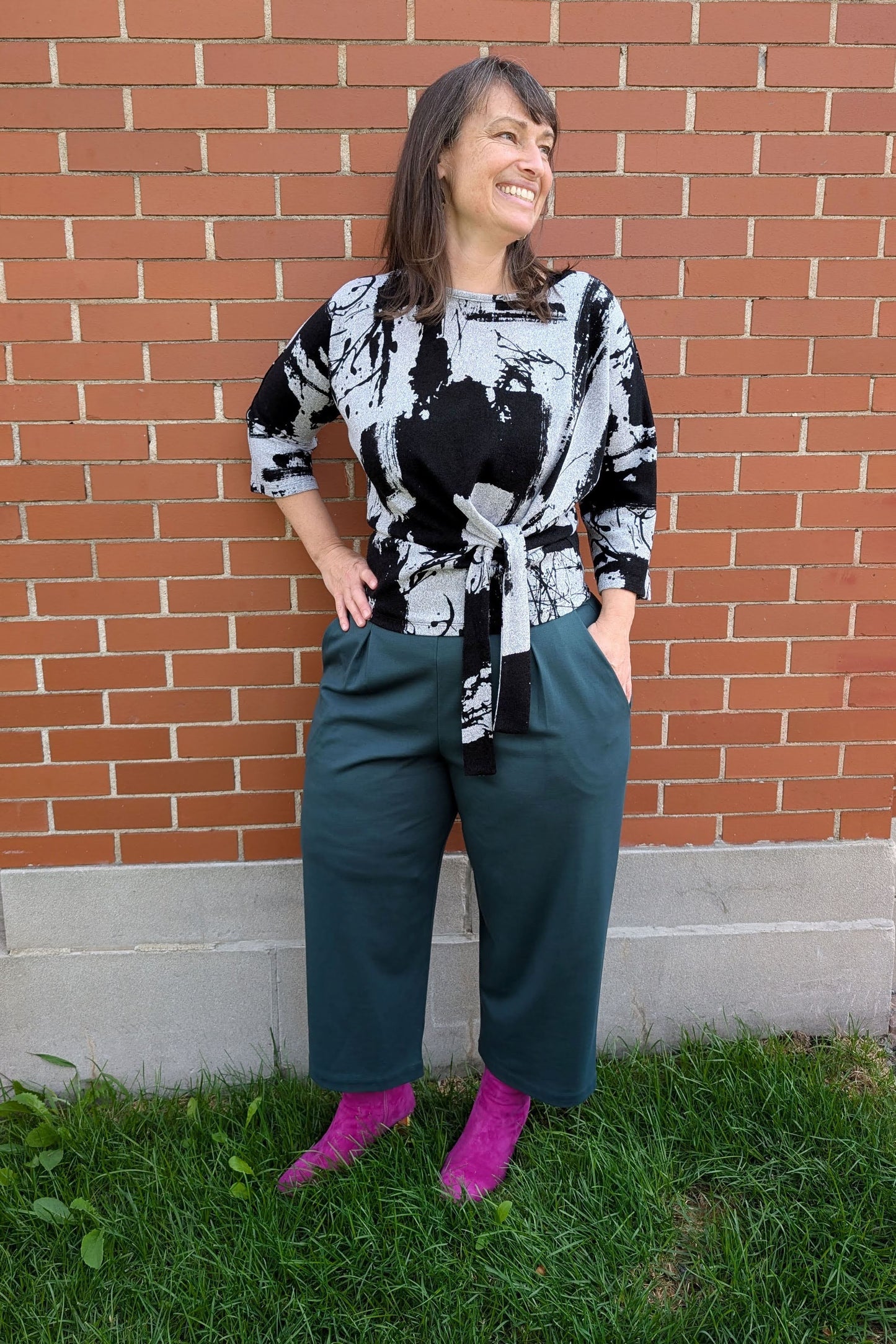 A woman wearing the NIna Top by Compli K with the Wilfred Pants by Melow in Forest - featuring
a wide pull-on waistband, front pleats, and cropped legs, standing in front of a brick wall.