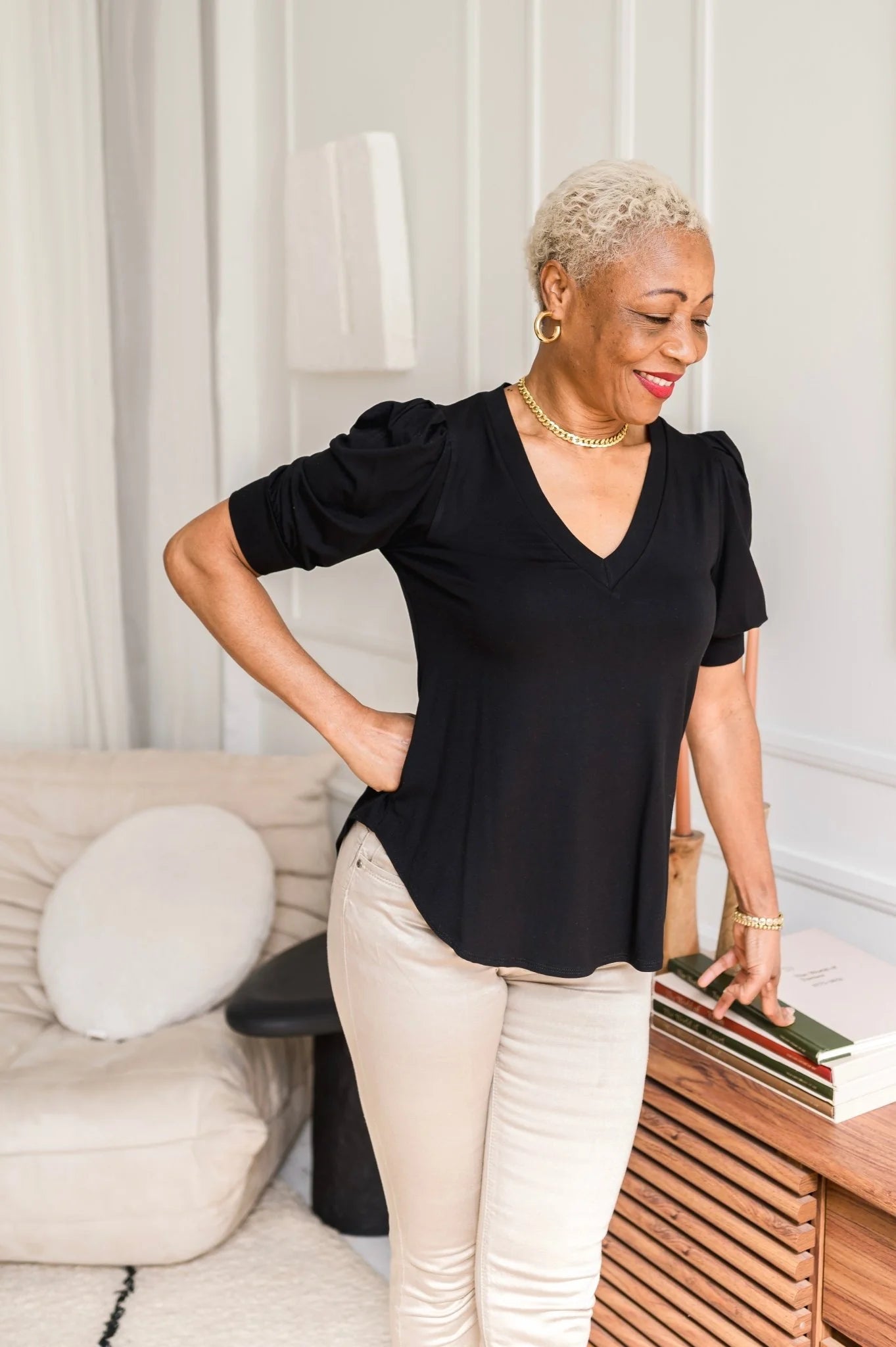 A woman wearing the Darling Tee by Dotty in Black with a v-neck, pleats at the shoulders, elbow-length sleeves, and a rounded hem. She is wearing it with white jeans and is standing in front of a white wall.
