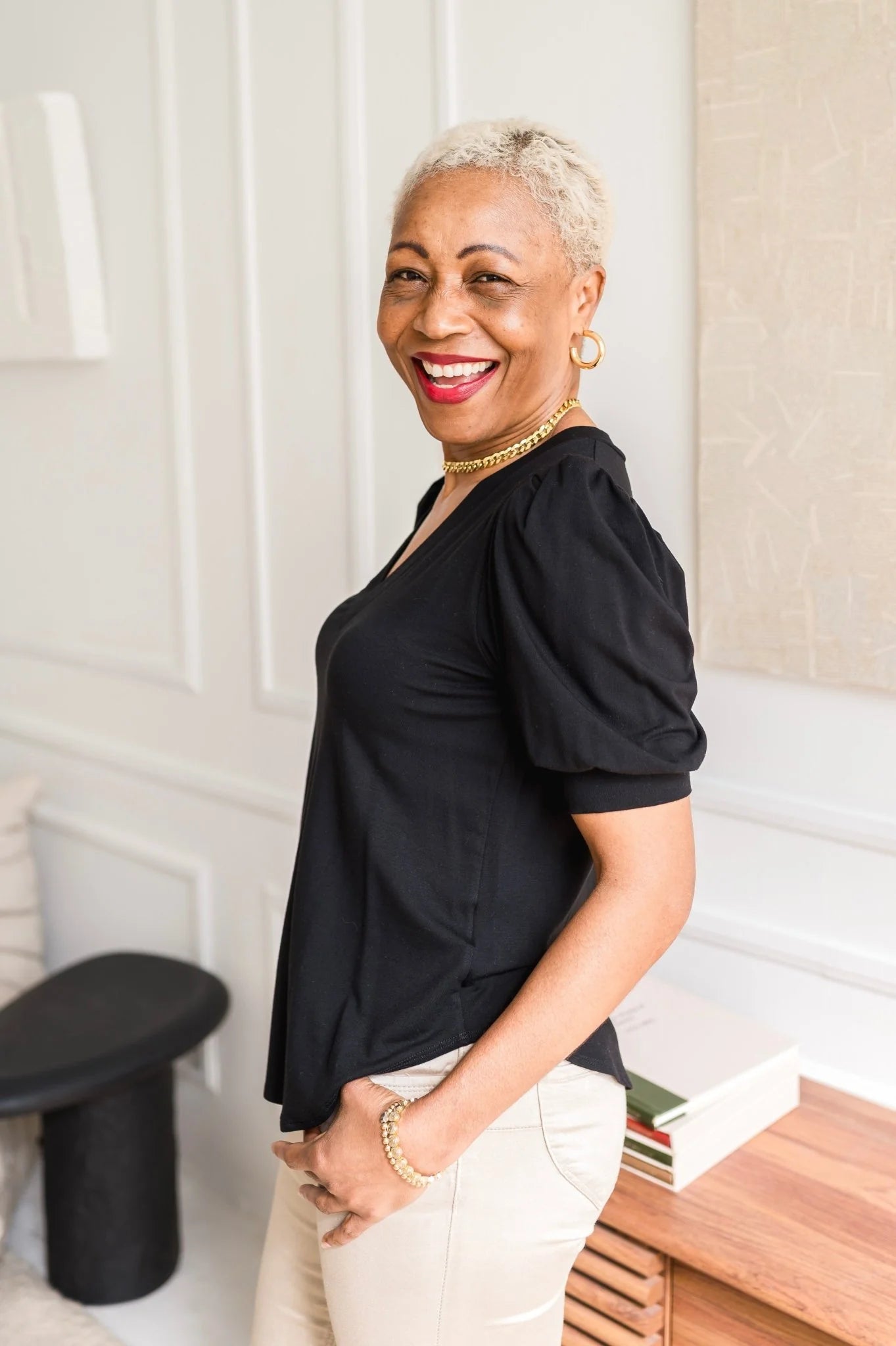 A woman wearing the Darling Tee by Dotty in Black with a v-neck, pleats at the shoulders, elbow-length sleeves, and a rounded hem. She is wearing it with white jeans and is standing in front of a white wall.