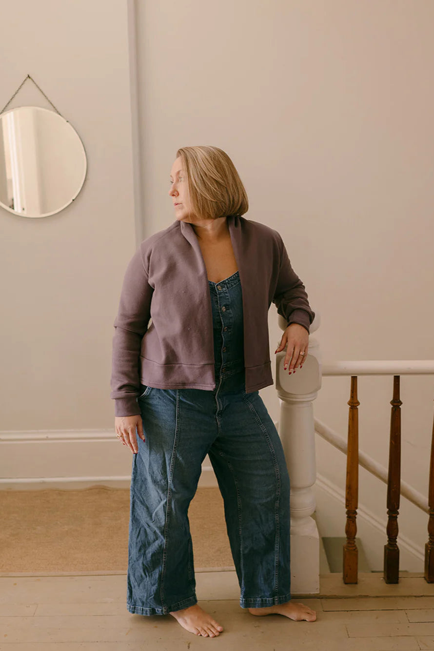 A woman wearing the Classic Spruce Cardigan by Blondie in Amethyst, a short cardigan with a subtle lapel collar, long sleeves, a wide ribbed hem. She is wearing it over denim overalls and is standing in front of a banister.