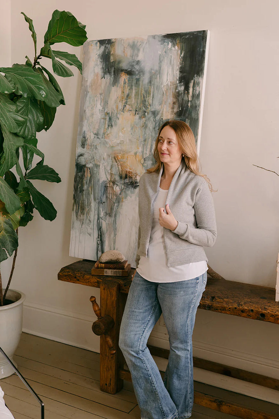 A woman wearing the Classic Spruce Cardigan by Blondie in Heather Grey, a short cardigan with a subtle lapel collar, long sleeves, a wide ribbed hem. She is wearing it with a white tee and jeans and is standing in front of a bench with a painting on it.