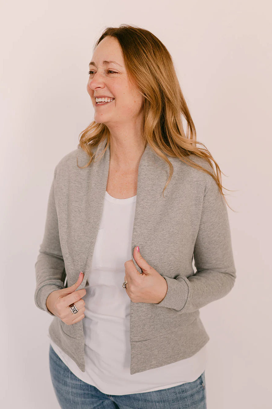 A woman wearing the Classic Spruce Cardigan by Blondie in Heather Grey, a short cardigan with a subtle lapel collar, long sleeves, a wide ribbed hem. She is wearing it with a white tee and jeans and is standing in front of a white background.