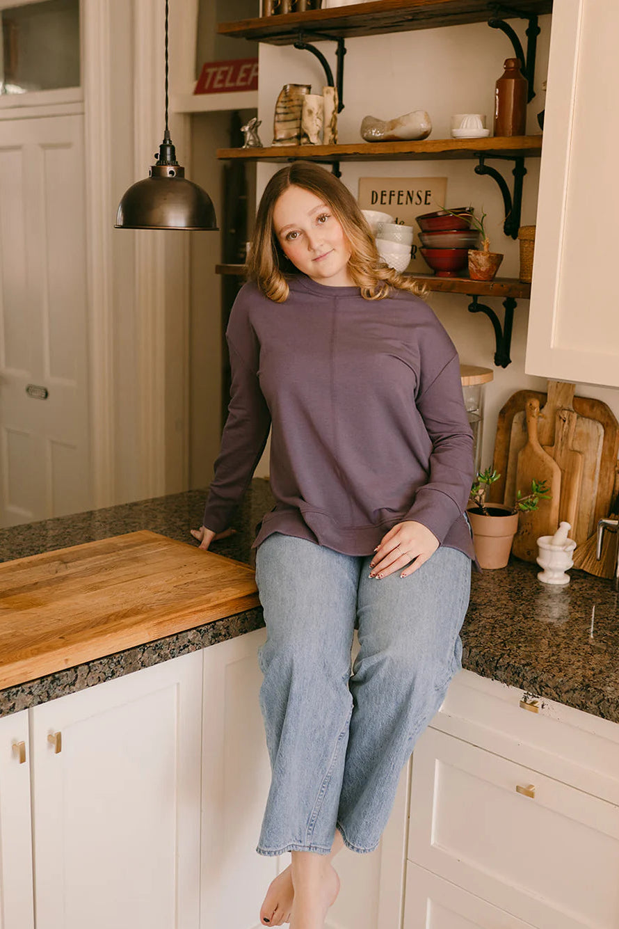 A woman wearing the Wheat Sweater by Blondie in Amethyst, with a crew neck, long dropped sleeves, a ribbed hem at the cuff, centre seams back and front, and small side slits. She is wearing it with jeans and sitting on a kitchen counter.