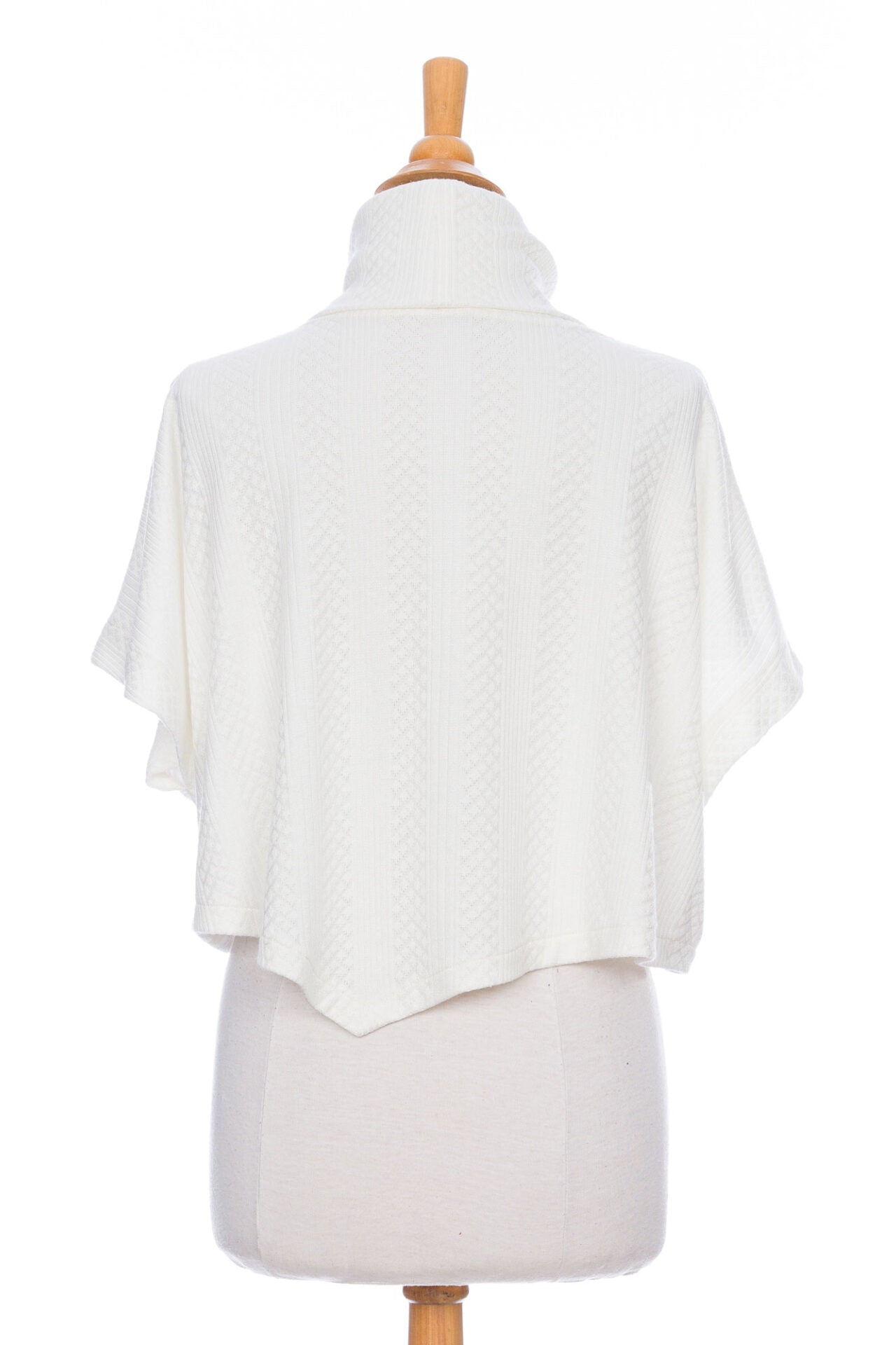 A back view of the Lupin Poncho-Bolero in Ivory by Rien ne se Perd, featuring soft jacquard knit, a loose turtleneck, and an asymmetrical hem, is shown on a mannequin in front of a white background.