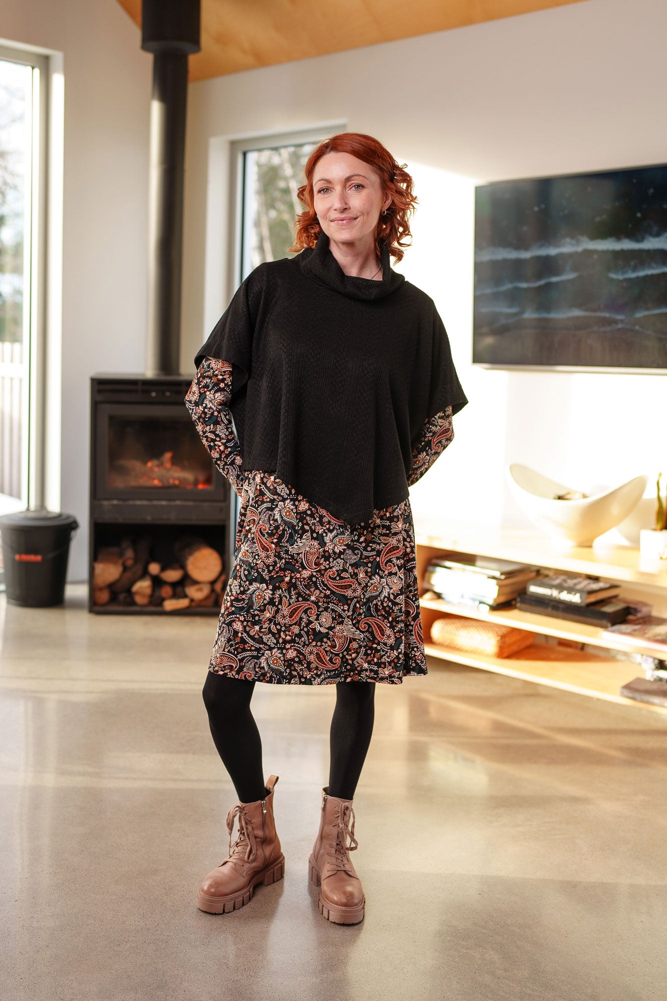 A woman wearing the Epilobe Dress by Rien ne se Perd in Fall Paisley, a classic wrap dress with a tie at the waist, delicate pleats at the shoulders, and a hem that hits above the knee. She is wearing the Lupin Poncho-Bolero in Black over it and is standing in a living room with a fireplace and TV.