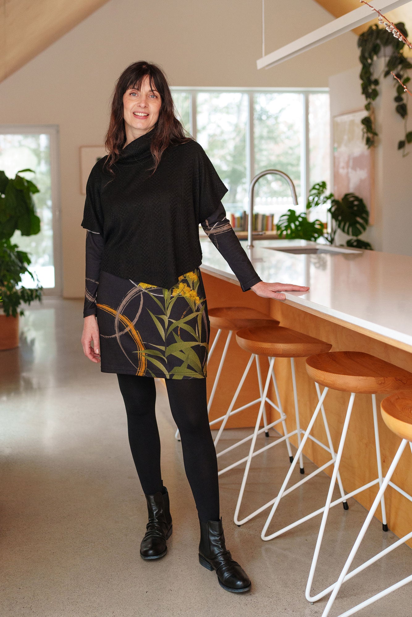 A woman wearing the Flore Dress by Rien ne se Perd in Bordure, a semi-fitted black dress with a flora inspired print, long sleeves, a round neck, and hem that hits above the knees. She is wearing it with the Lupin Poncho-Bolero in Black and is standing in a kitchen.