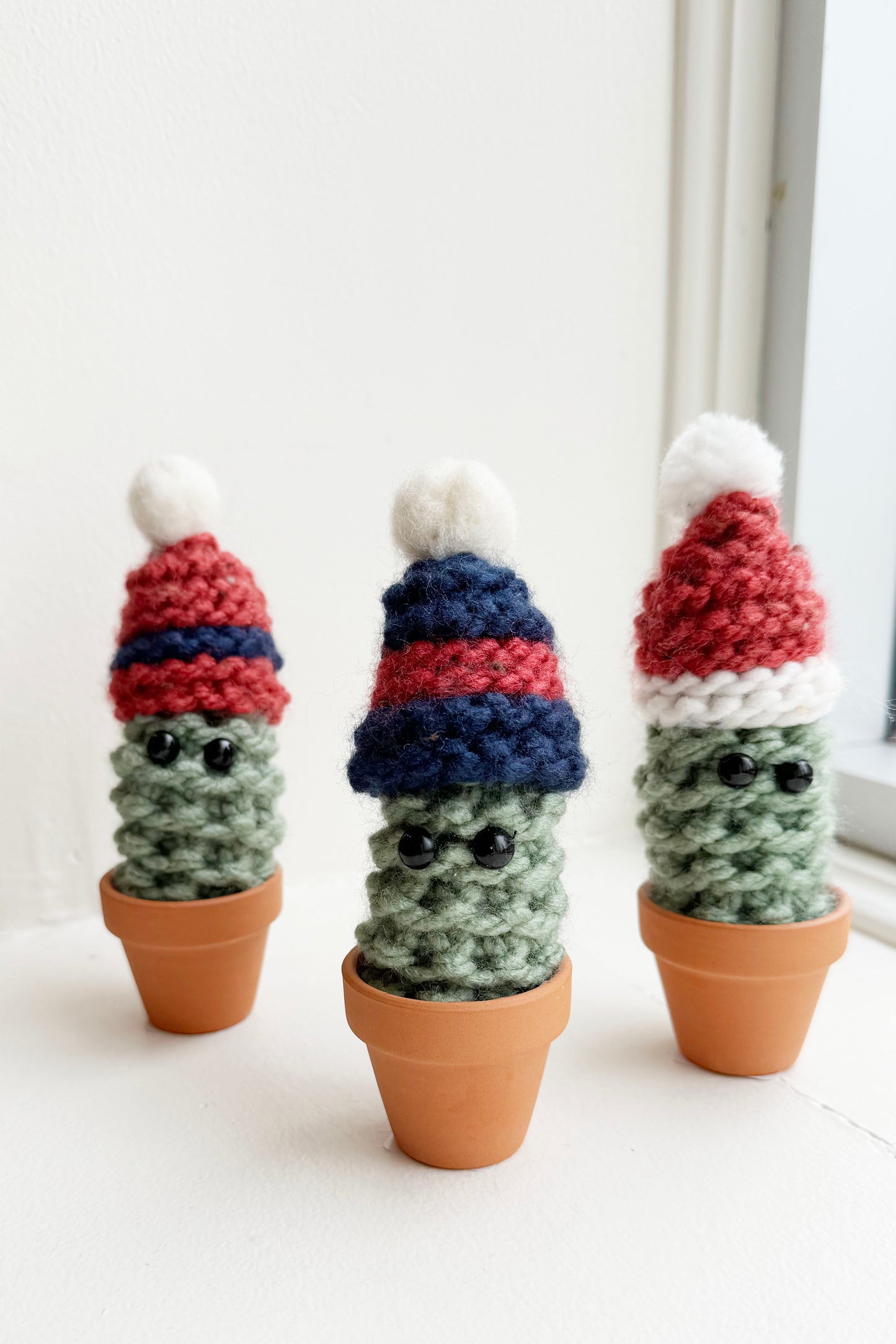 Knitted Winter Cacti by Curl Country, with hats and set in terracotta pots, are shown in front of a white background.