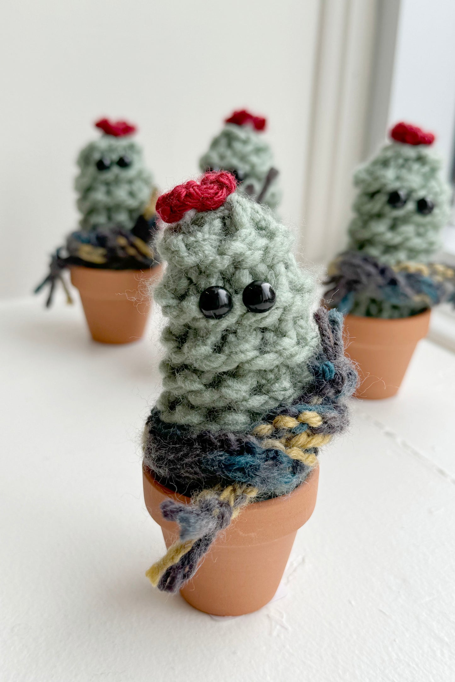 Knitted Winter Cacti by Curl Country, with scarves and set in terracotta pots, are shown in front of a white background.