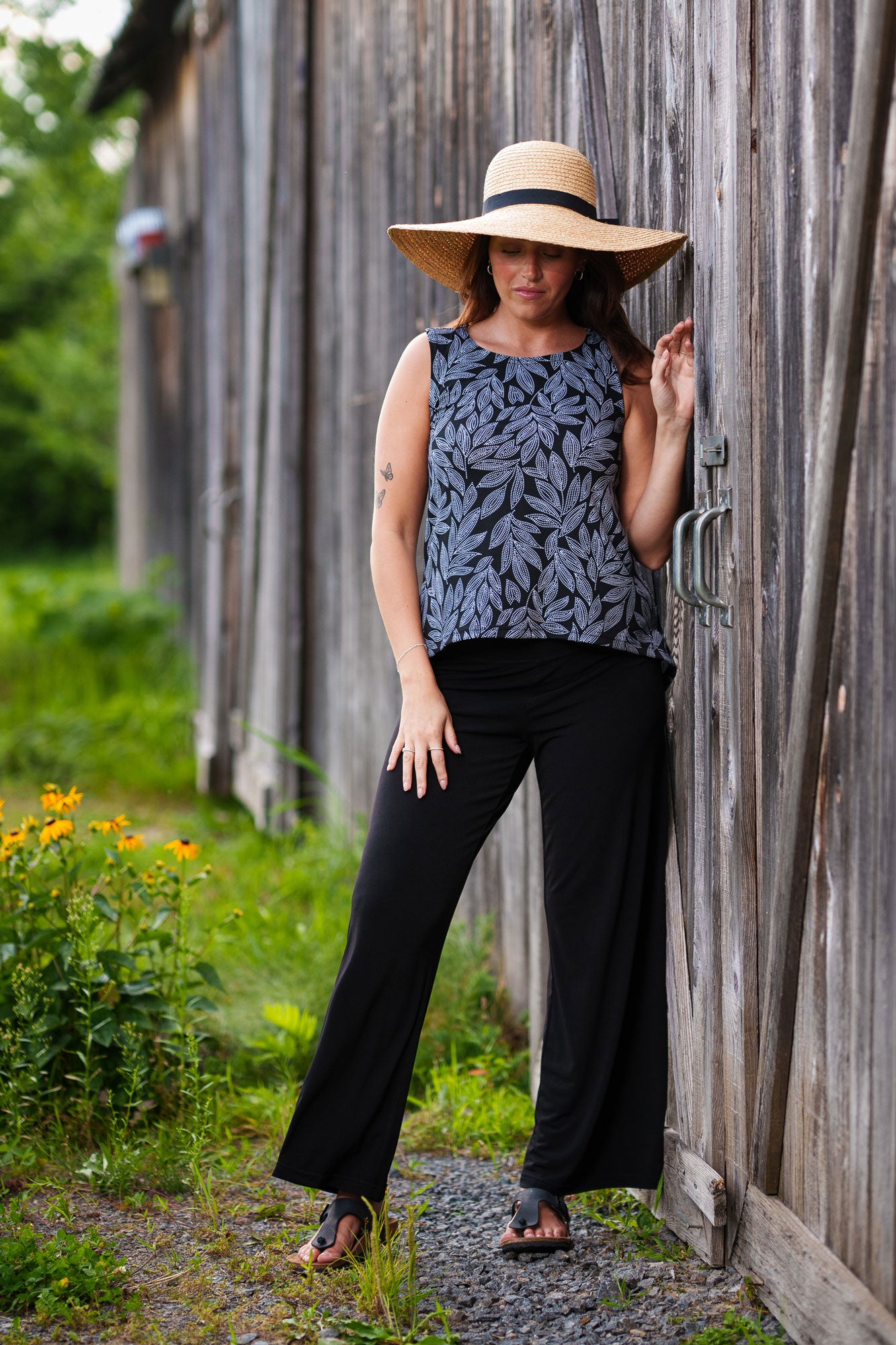 A woman wearing the Rumba Palazzo Pants in Black and the Swing Cami in Black and White Foliage by Rien ne se Perd, standing outside in front of a wooden building.