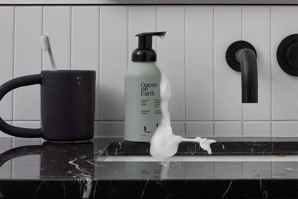 Pump bottle of Guests on Earth Foaming Hand Soap in Woodlands scent, on a bathroom counter next to a black mug and toothbrush.