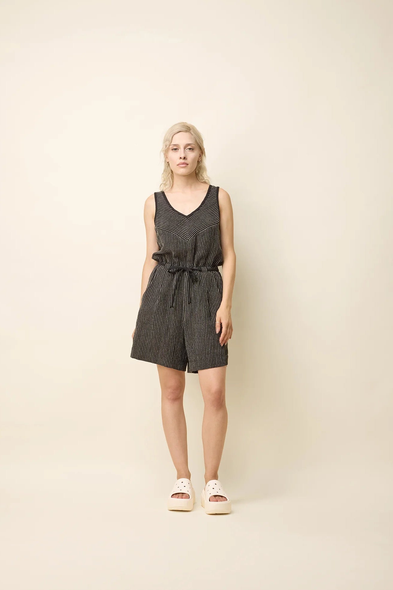 A woman wearing the Shamsa Romper by Cokluch in Black Stripe, a sleeveless romper with a V-neck at the front, a cross-over back, and elastic waist with a tie belt. She is standing in front of a beige background.