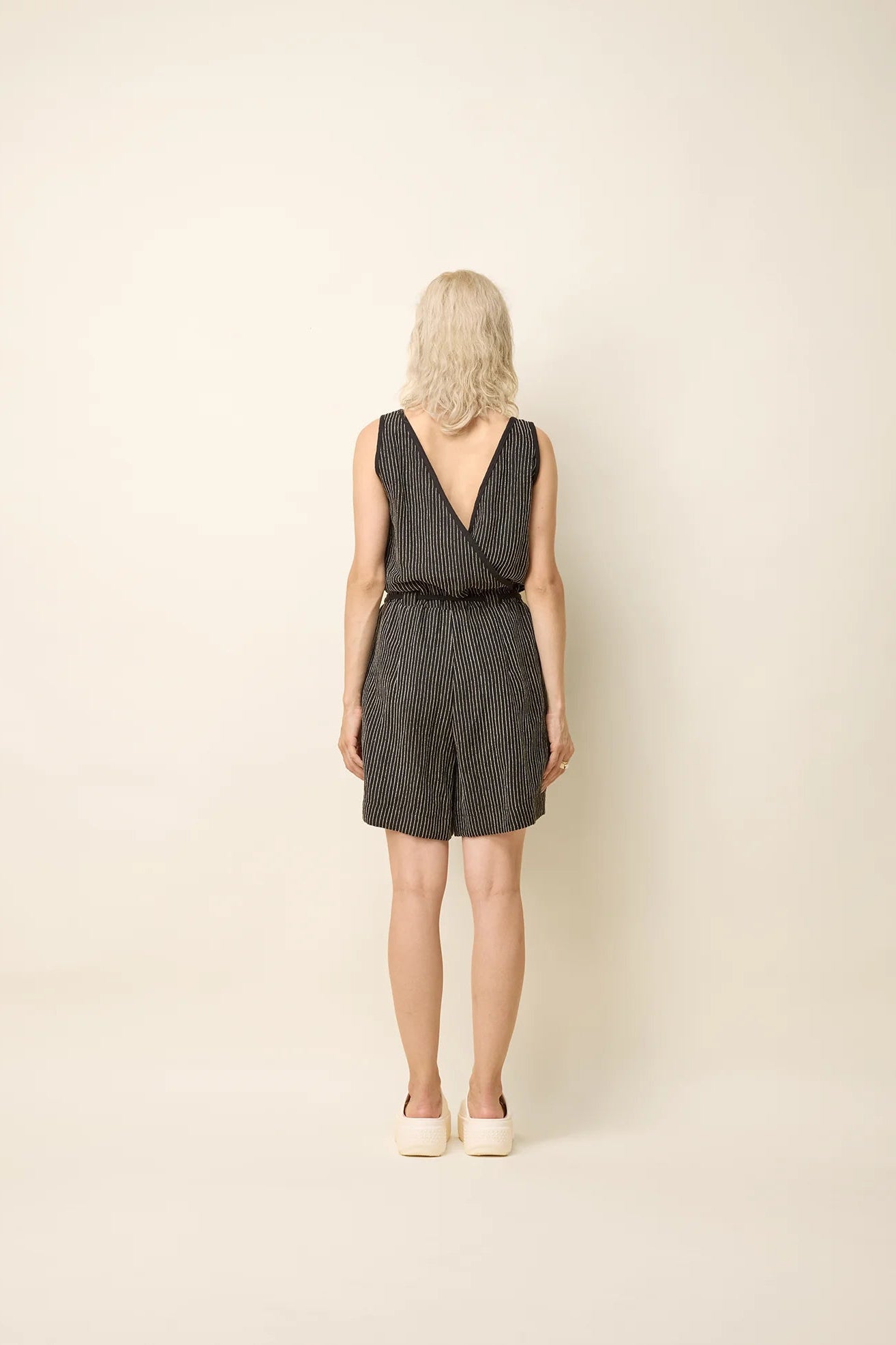 Back view of a woman wearing the Shamsa Romper by Cokluch in Black Stripe, a sleeveless romper with a V-neck at the front, a cross-over back, and elastic waist with a tie belt. She is standing in front of a beige background.