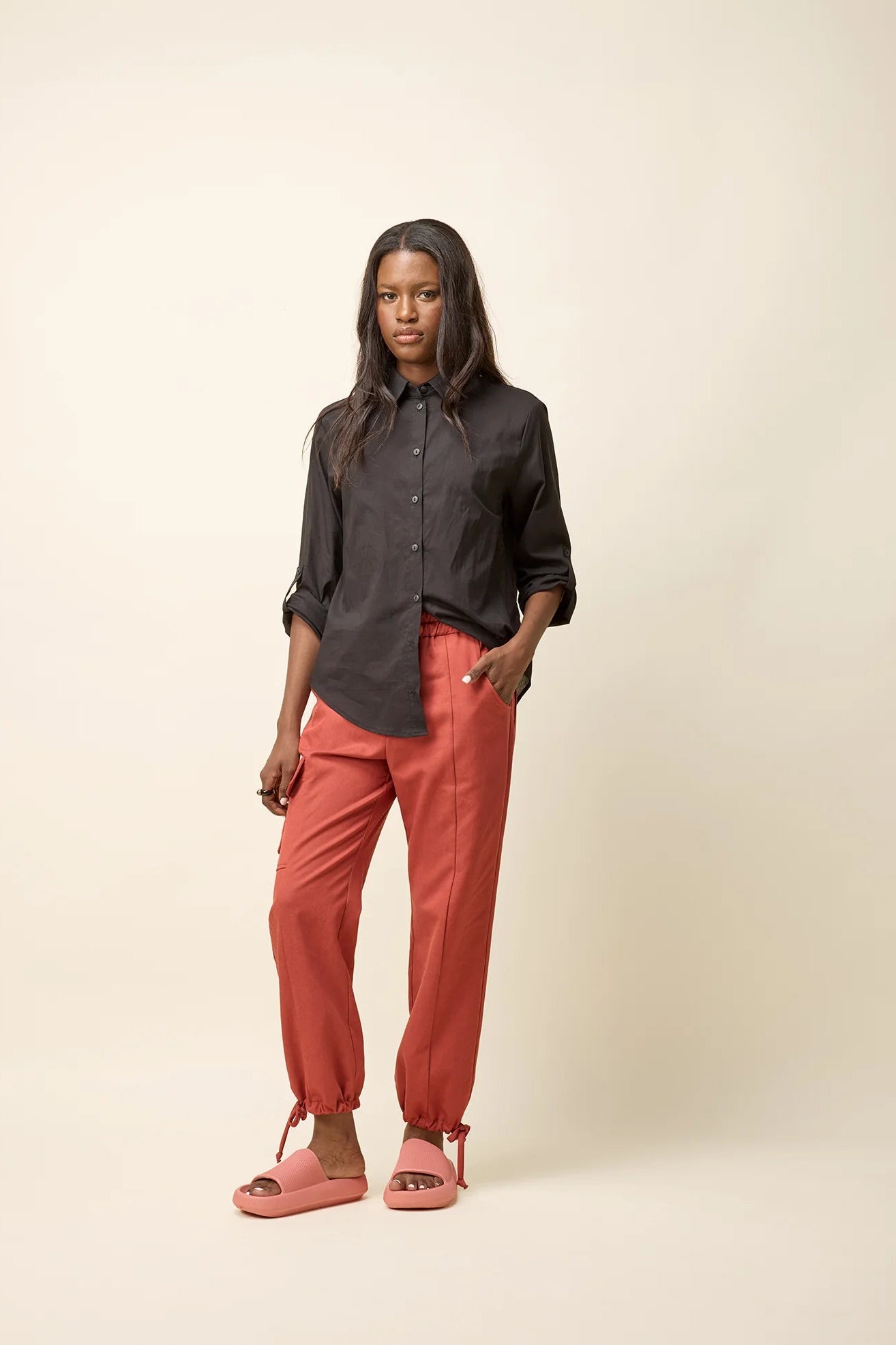 A woman wearing the Chila Shirt by Cokluch in Black, a button down shirt with a blouse collar, rounded hem that falls below the hip, and long sleeves that can be cinched with a button and strap closure. She is wearing it with orange pants and standing in front of a white background.