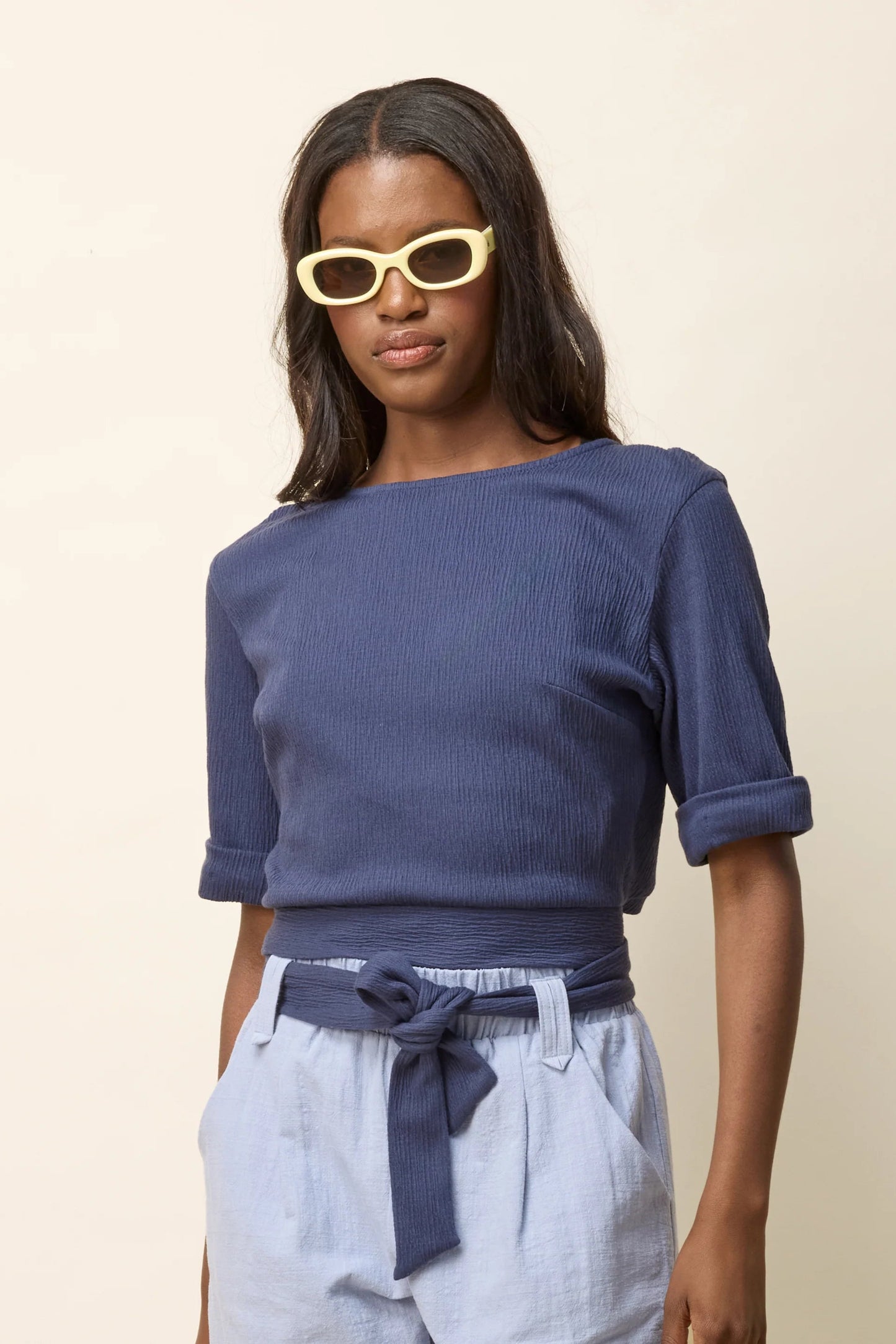 3/4 view of a woman wearing the Jess Top by Cokluch in Navy, a fitted top with a round neck, cuffed elbow-length sleeves, a cut out at the bottom back, and a tie belt. She is wearing it with light blue pants and standing in front of a beige background.