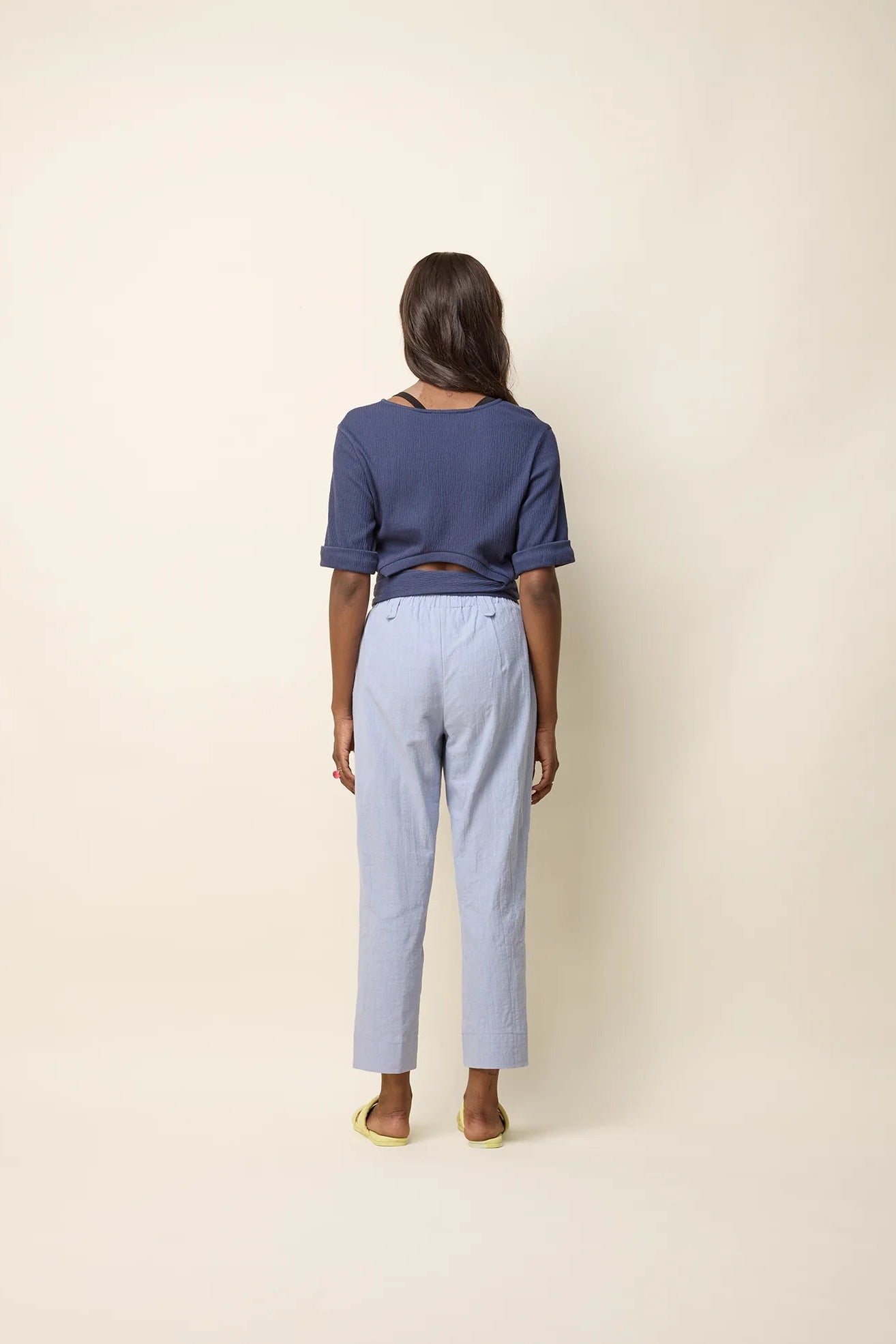 A woman wearing the Jess Top by Cokluch in Navy, a fitted top with a round neck, cuffed elbow-length sleeves, a cut out at the bottom back, and a tie belt. She is wearing it with light blue pants and standing in front of a beige background.