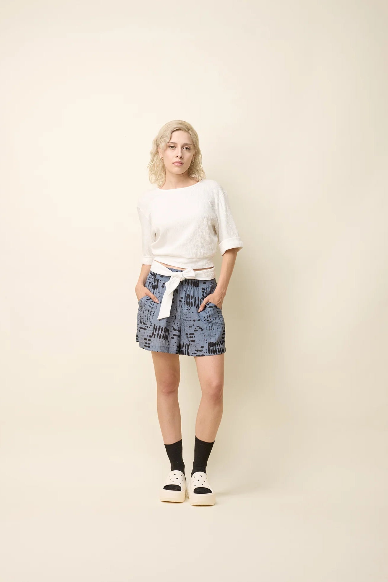 A woman wearing the Jess Top by Cokluch in Off-White, a fitted top with a round neck, cuffed elbow-length sleeves, a cut out at the bottom back, and a tie belt. She is wearing it with the Joy Shorts in Blue Ink and standing in front of a beige background.