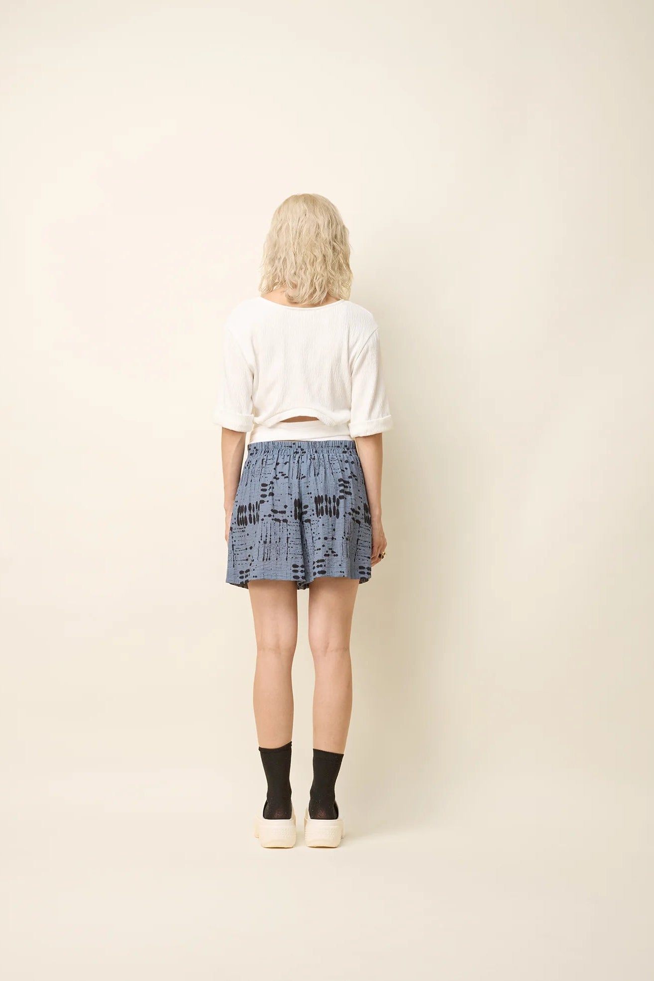 Back view of a woman wearing the Jess Top by Cokluch in Off-White, a fitted top with a round neck, cuffed elbow-length sleeves, a cut out at the bottom back, and a tie belt. She is wearing it with the Joy Shorts in Blue Ink and standing in front of a beige background.